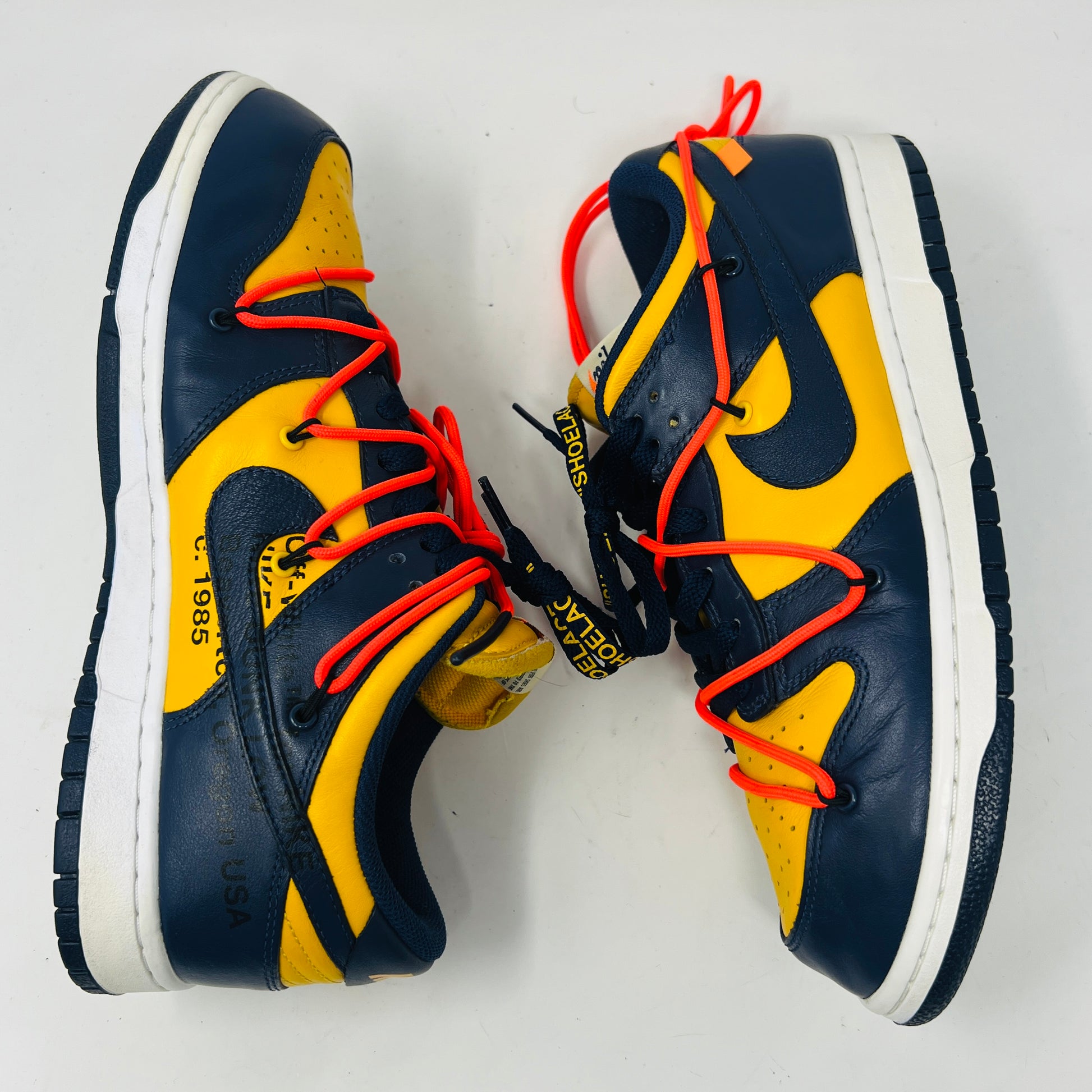 The 2019 Nike Dunk Low Off-White University Gold sneakers feature a navy and yellow design, accented with bold red laces and attached white tags and a zip tie. Displayed atop their box, the label lists them as US size 10.5 and UK size 12, all set against a plain white background.