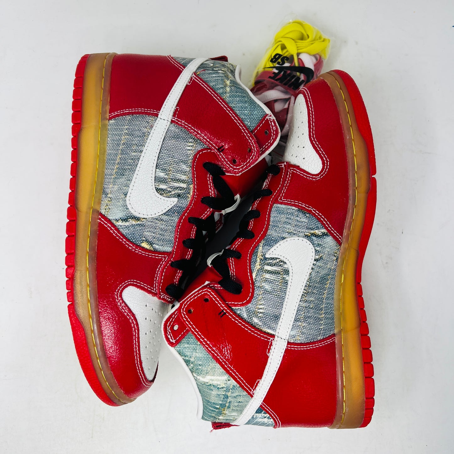 The Nike SB Dunk High Shoe Goo from 2008, priced at $525, sits atop its box. It features red and blue denim sections, a white swoosh, and a gum sole. A tag shows size and numbers, with extra laces beside the shoebox.
