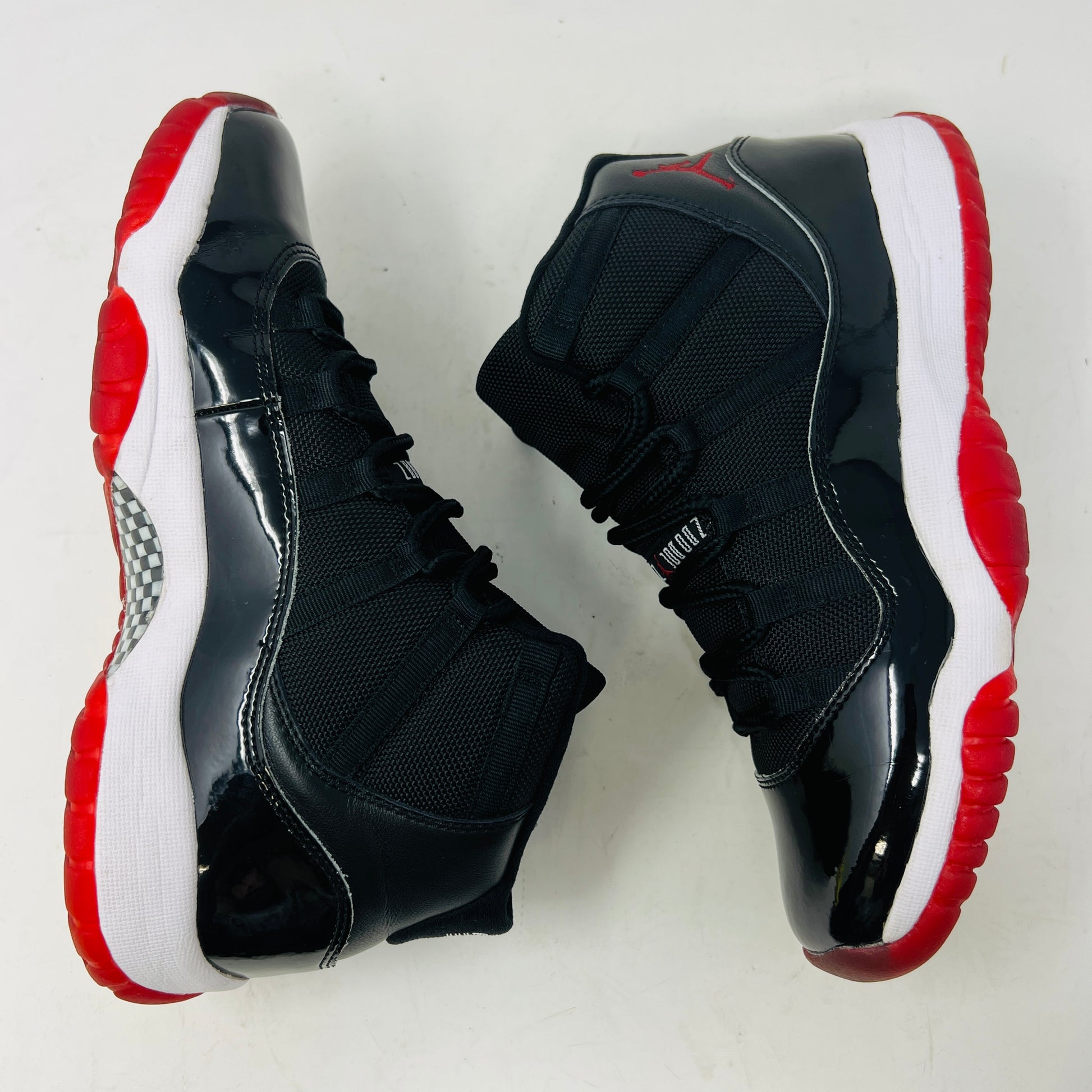 A pristine black and red Jordan 11 Retro Playoffs Bred (2019) (GS) sneaker is on an orange Nike box, featuring a patent leather upper, white midsole, red outsole, and the Jumpman logo near the heel. The label indicates year and size details.