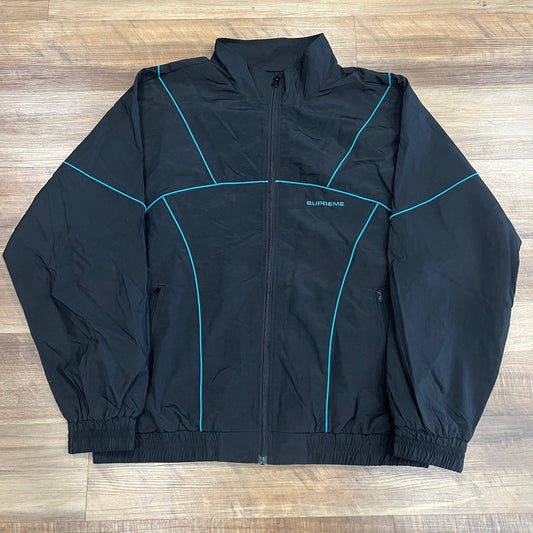 The Supreme Piping Track Jacket (SS19) in black features teal piping, elastic cuffs, a zip-up front, and a subtle logo on the left chest. The extra-large jacket is displayed laid flat on a wooden floor.
