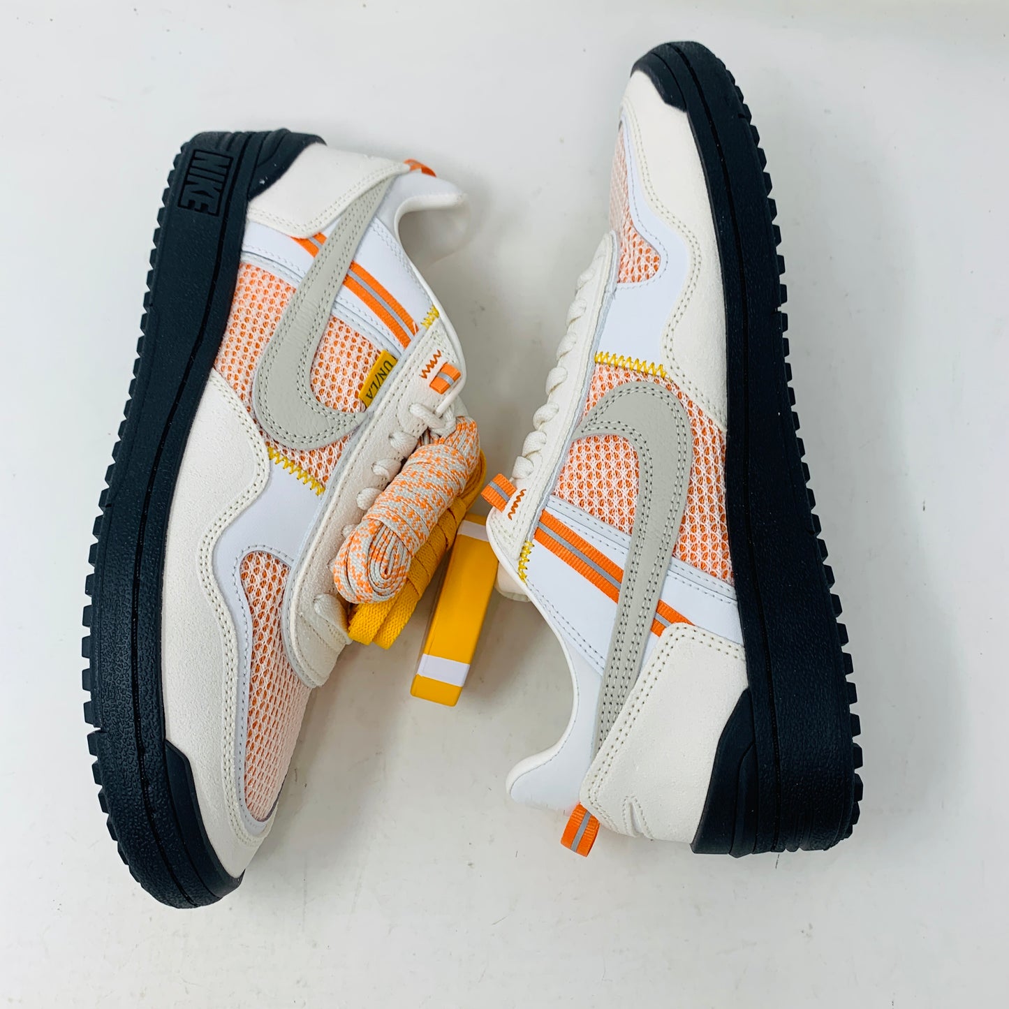 The pristine Nike Field General Union LA Ivory sneakers, in white and orange with black soles, rest on an orange box. Featuring a UNION tag, Nike logo, and a blue sticker with a silhouetted man, theyre perfect for any accessories collection.