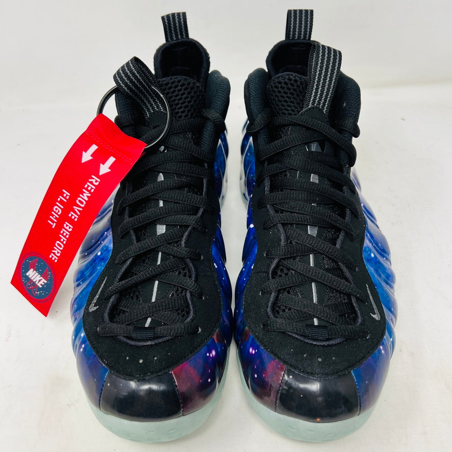 The Nike Air Foamposite One Galaxy (2025) sneaker, showcasing vivid blues, purples, and reds in a starry design, sits atop a pristine black box. The translucent icy blue sole adds a dazzling touch.