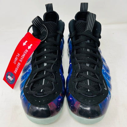 The Nike Air Foamposite One Galaxy (2025) sneaker, showcasing vivid blues, purples, and reds in a starry design, sits atop a pristine black box. The translucent icy blue sole adds a dazzling touch.