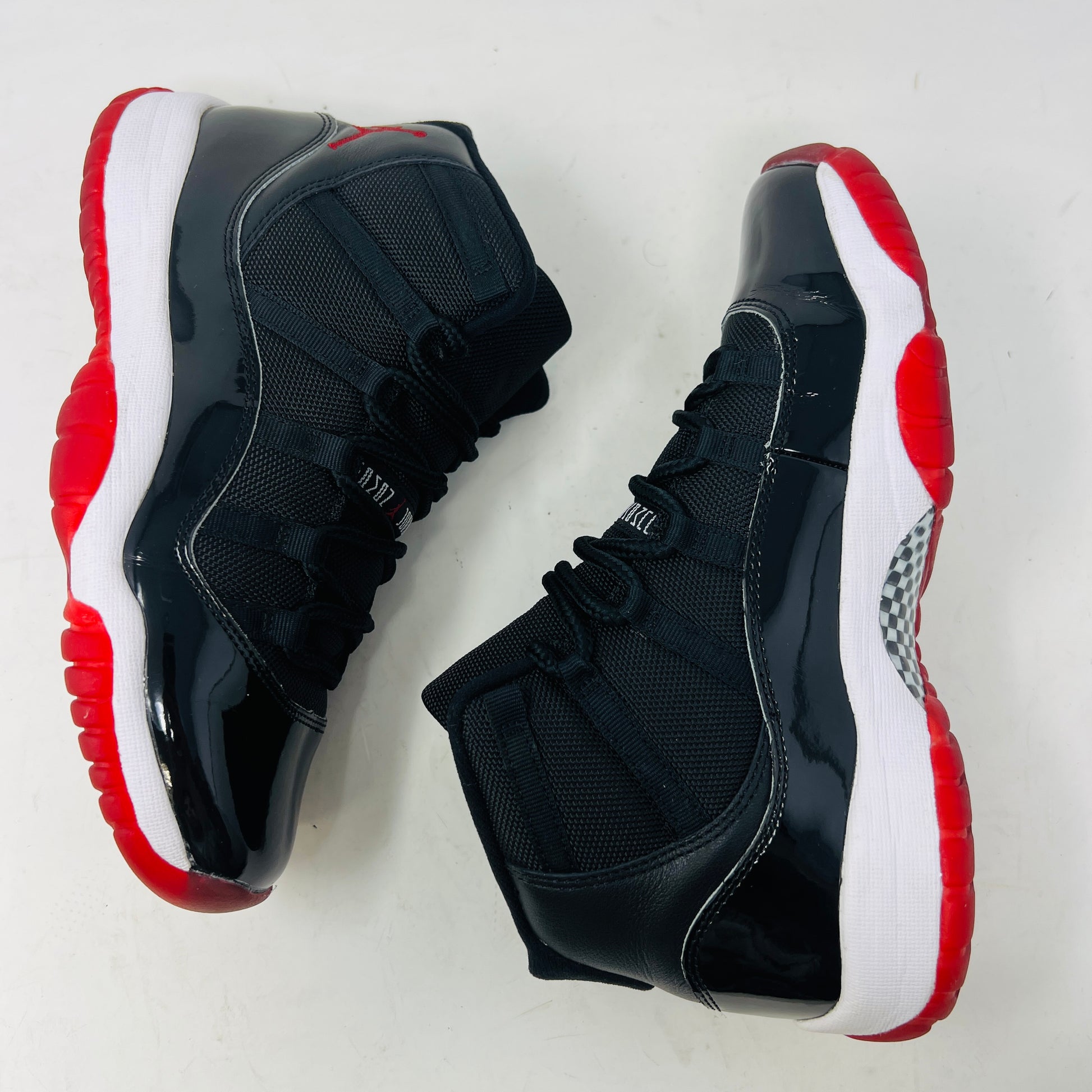 A pristine black and red Jordan 11 Retro Playoffs Bred (2019) (GS) sneaker is on an orange Nike box, featuring a patent leather upper, white midsole, red outsole, and the Jumpman logo near the heel. The label indicates year and size details.