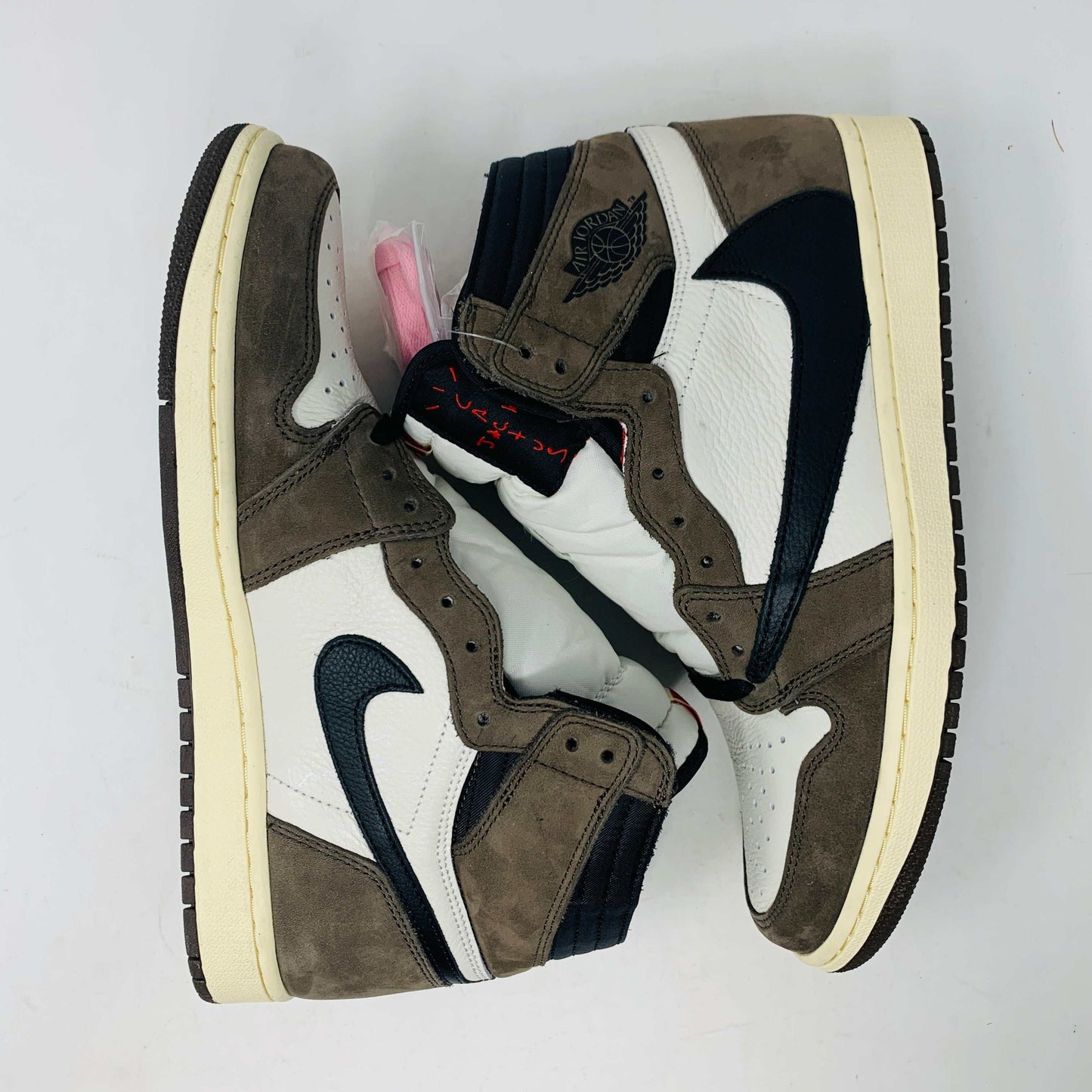 A pair of authentic Jordan 1 High Travis Scott Mocha sneakers in brown, white, and black is displayed on an orange and brown Nike shoebox. These feature a distinctive reversed black Swoosh and come with extra laces for versatile styling.