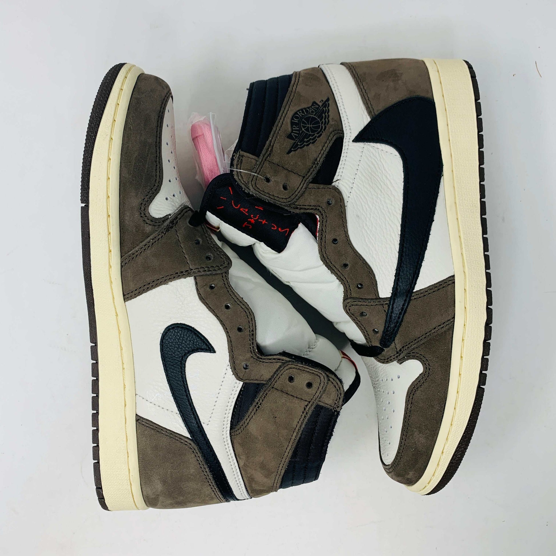 A pair of authentic Jordan 1 High Travis Scott Mocha sneakers in brown, white, and black is displayed on an orange and brown Nike shoebox. These feature a distinctive reversed black Swoosh and come with extra laces for versatile styling.