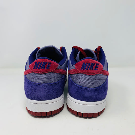 A pair of Nike Dunk Low Plum sneakers, in purple and maroon, rests on a red shoe box featuring a white Nike logo on a plain background. The shoes showcase a red swoosh and white midsoles, confirming their authenticity from Nike.