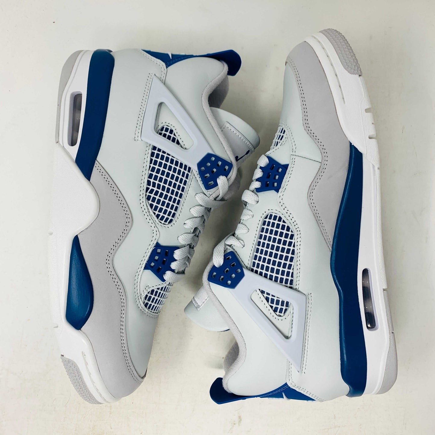 The Jordan 4 Retro Military Blue (2024) sneaker, with white and blue mesh details and a visible air cushion, rests on a black speckled NIKE AIR box with bold orange text. It is 100% authentic, including orange tags, ensuring it’s brand new.
