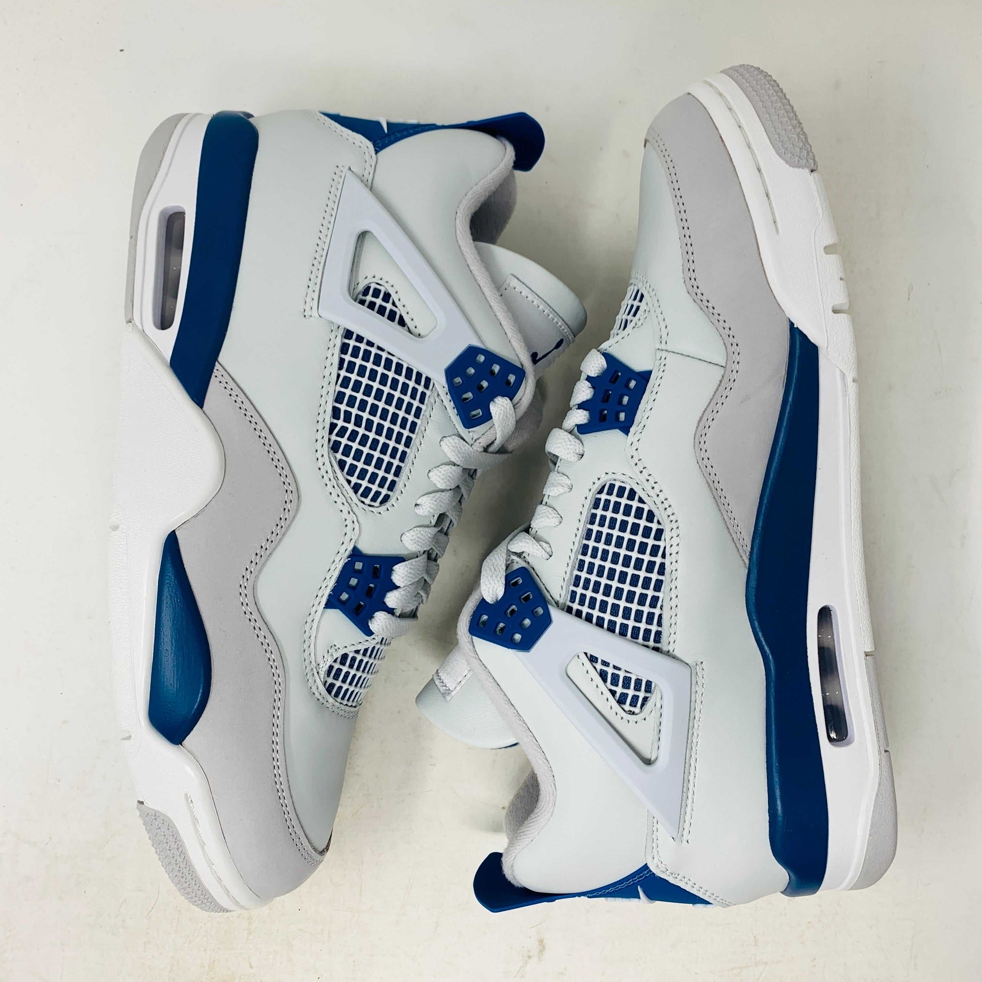 The Jordan 4 Retro Military Blue (2024) sneaker, with white and blue mesh details and a visible air cushion, rests on a black speckled NIKE AIR box with bold orange text. It is 100% authentic, including orange tags, ensuring it’s brand new.
