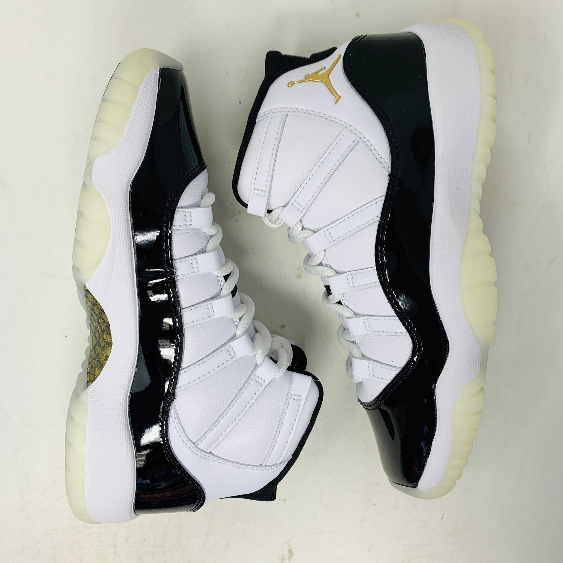 A pair of authentic Jordan 11 Retro DMP Gratitude (2023) (GS) sneakers, featuring a glossy white and black finish with a gold logo, is placed on top of a gray Nike Air shoebox.