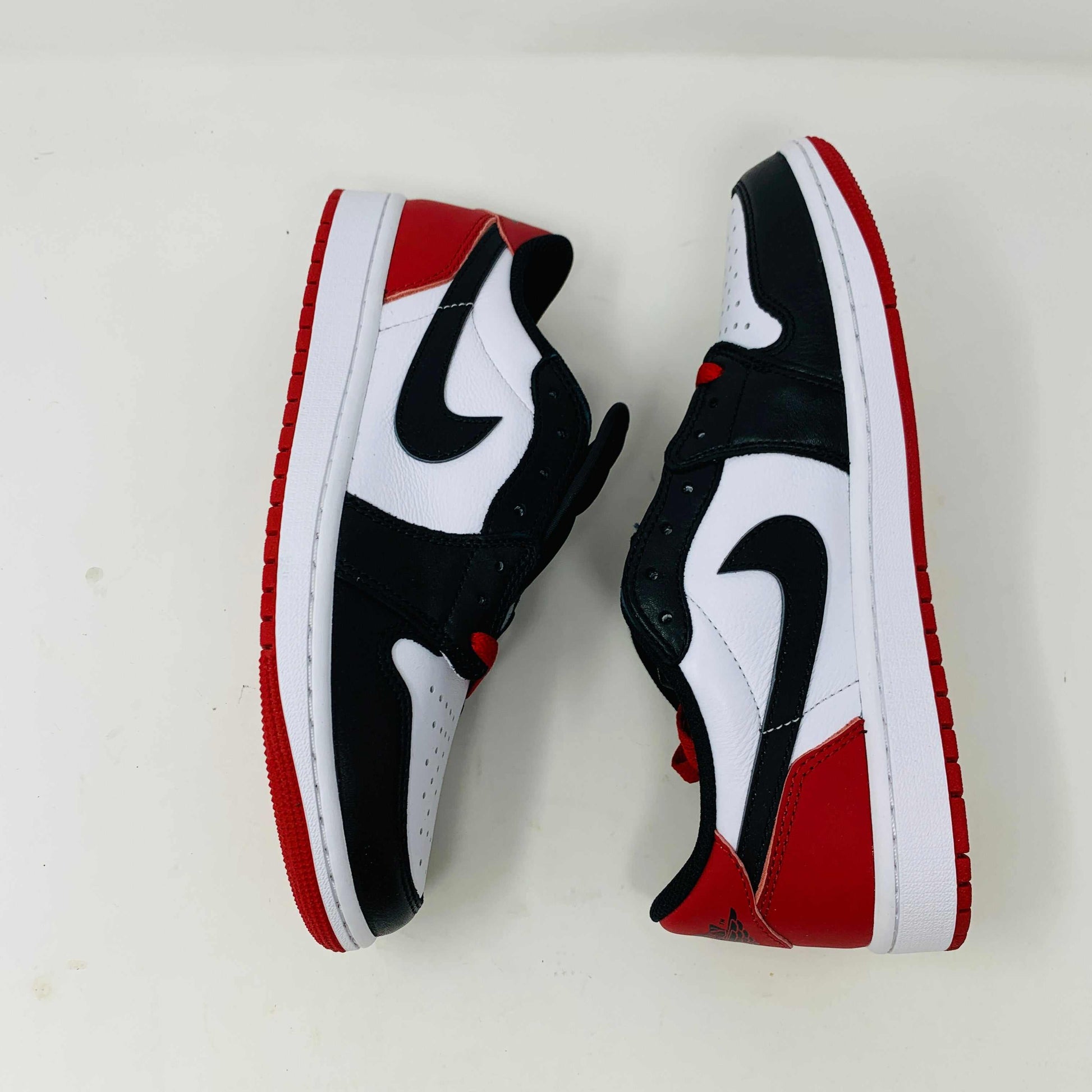 Discover the Jordan 1 Retro Low OG Black Toe (2023) sneakers featuring genuine black and white leather panels, red laces, and soles. These low-tops from Jordan come with extra laces and are displayed on a clean white surface against a pristine background.