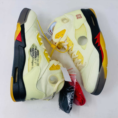 The Jordan 5 Off White Sail sneakers feature a yellow and black design with red accents, a visible air unit, and the number 23 on the side. BRAND NEW in a black shoebox with a red Jumpman logo and speckled base, they include an attached yellow tag and come with EXTRA LACES for versatility.