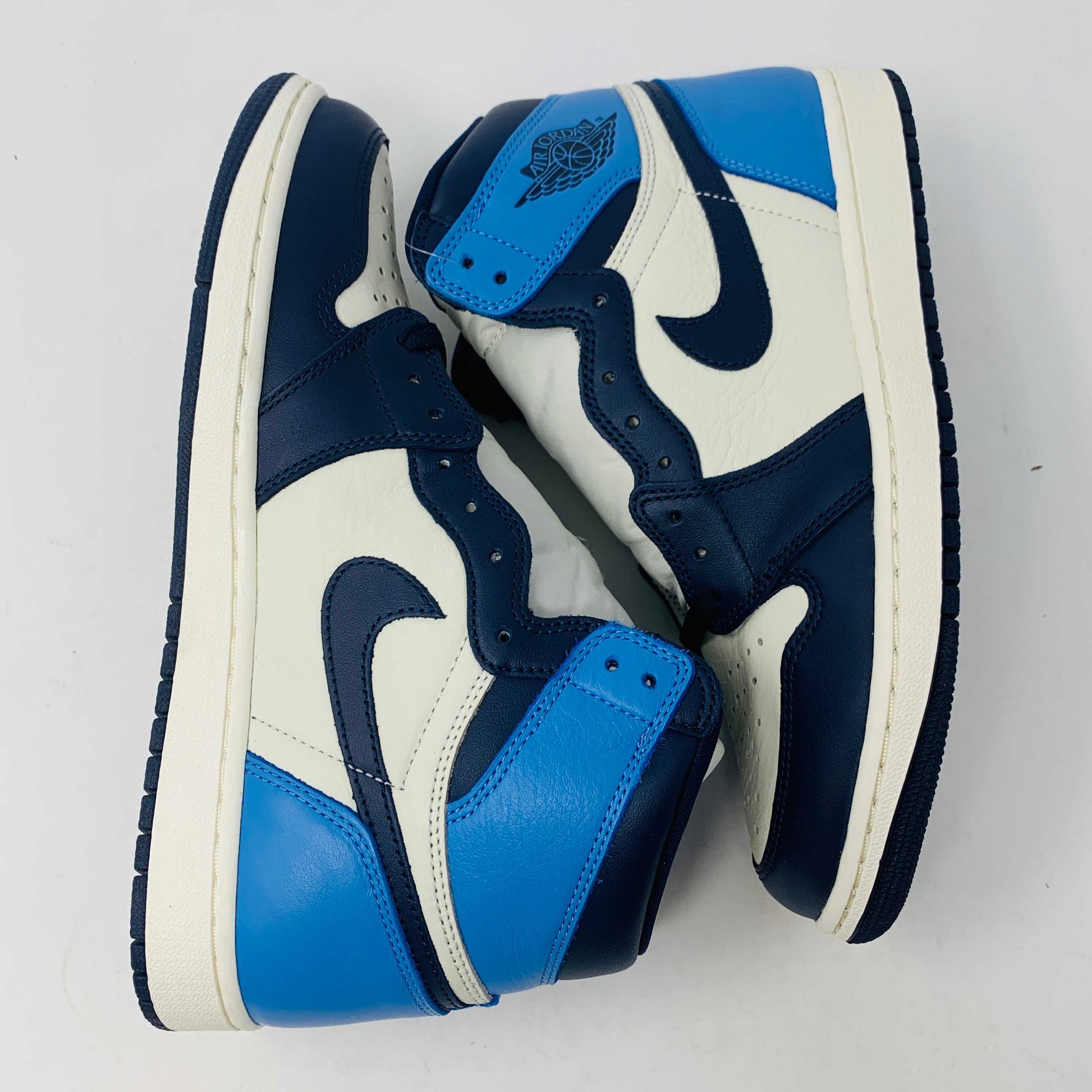 The new Jordan 1 Obsidian high-top sneaker, in blue and white with a black swoosh and blue laces, is displayed on a black and red box featuring 1985 Air Jordan 1 text. A printed Air Jordan wings logo is near the ankle.