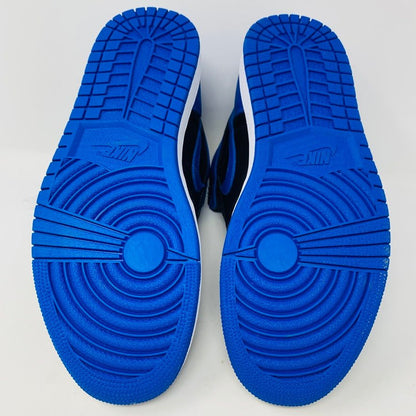 A pair of brand new black and blue Jordan 1 Royal 2017 high-tops are showcased on a black and red Nike box, with a small plastic bag holding authentic blue laces attached to one shoe.