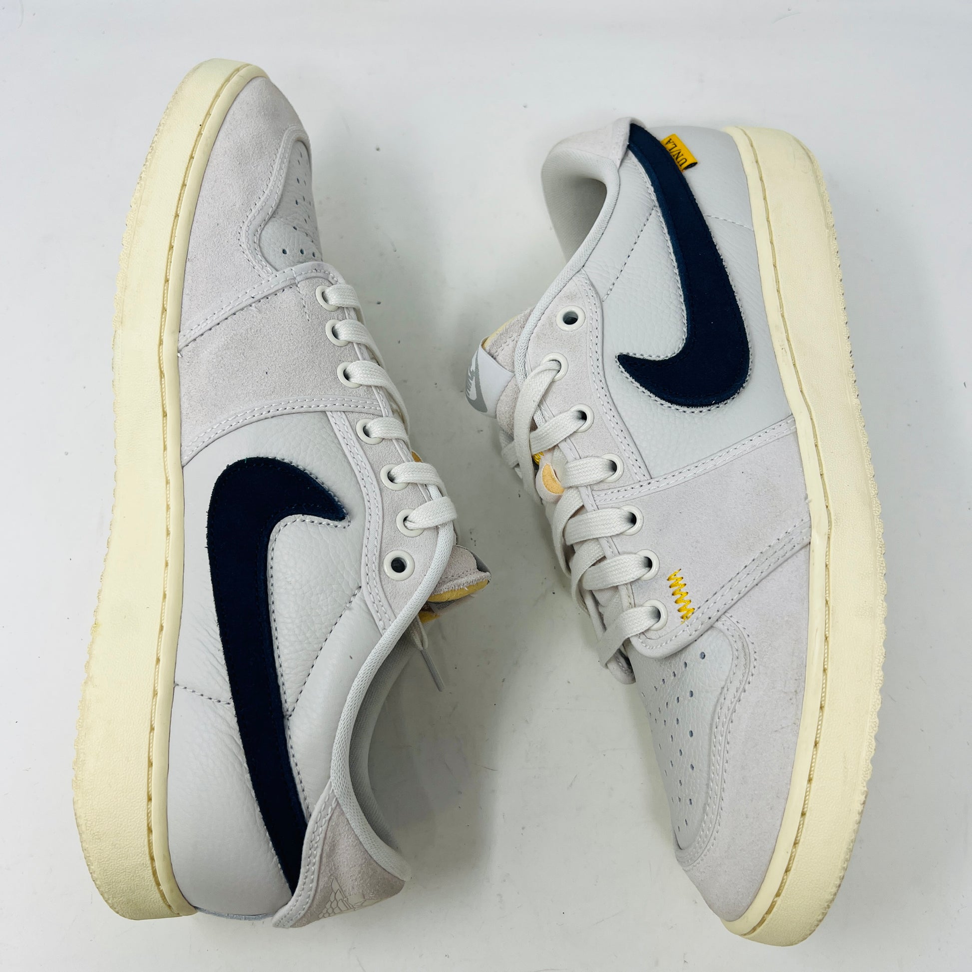 The Jordan 1 Retro AJKO Low SP Union Sail Leather sneakers feature black swooshes, subtle yellow stitching, and white laces. Pictured on a pristine white background, they include extra laces for versatile styling.