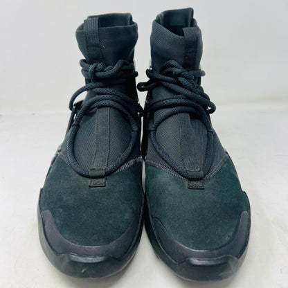 The Nike Air Fear of God 1 Triple Black is a high-top, dark, minimalist sneaker with angular overlays and a textured sole. It features a high collar, matte finish, dark laces, and has a small logo near the midsole. The background is light gray with general wear signs.