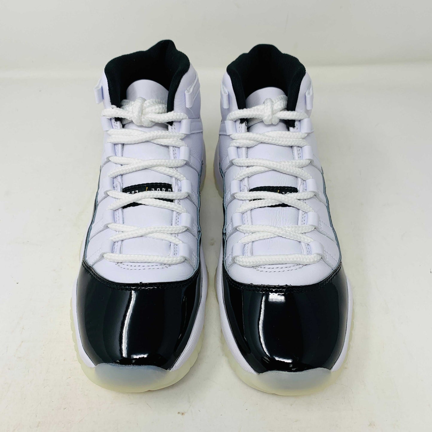A pair of authentic Jordan 11 Retro DMP Gratitude (2023) (GS) sneakers, featuring a glossy white and black finish with a gold logo, is placed on top of a gray Nike Air shoebox.
