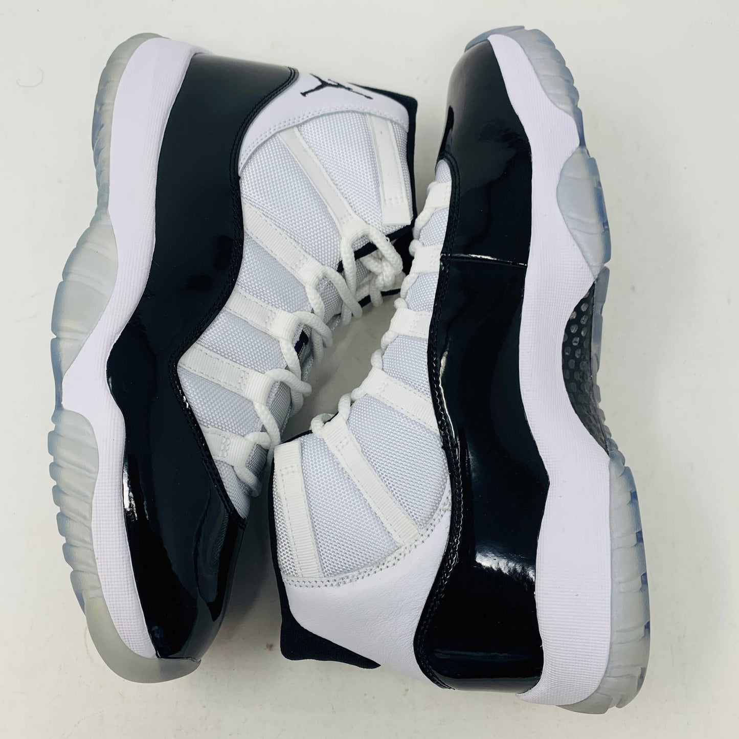 A BRAND NEW black and white Jordan 11 Concord 2018 sneaker sits on its shoebox, showcasing a glossy black patent leather finish and white mesh upper with the iconic Jumpman logo. The shoebox, bearing a white Nike Air logo, ensures these shoes are 100% authentic.