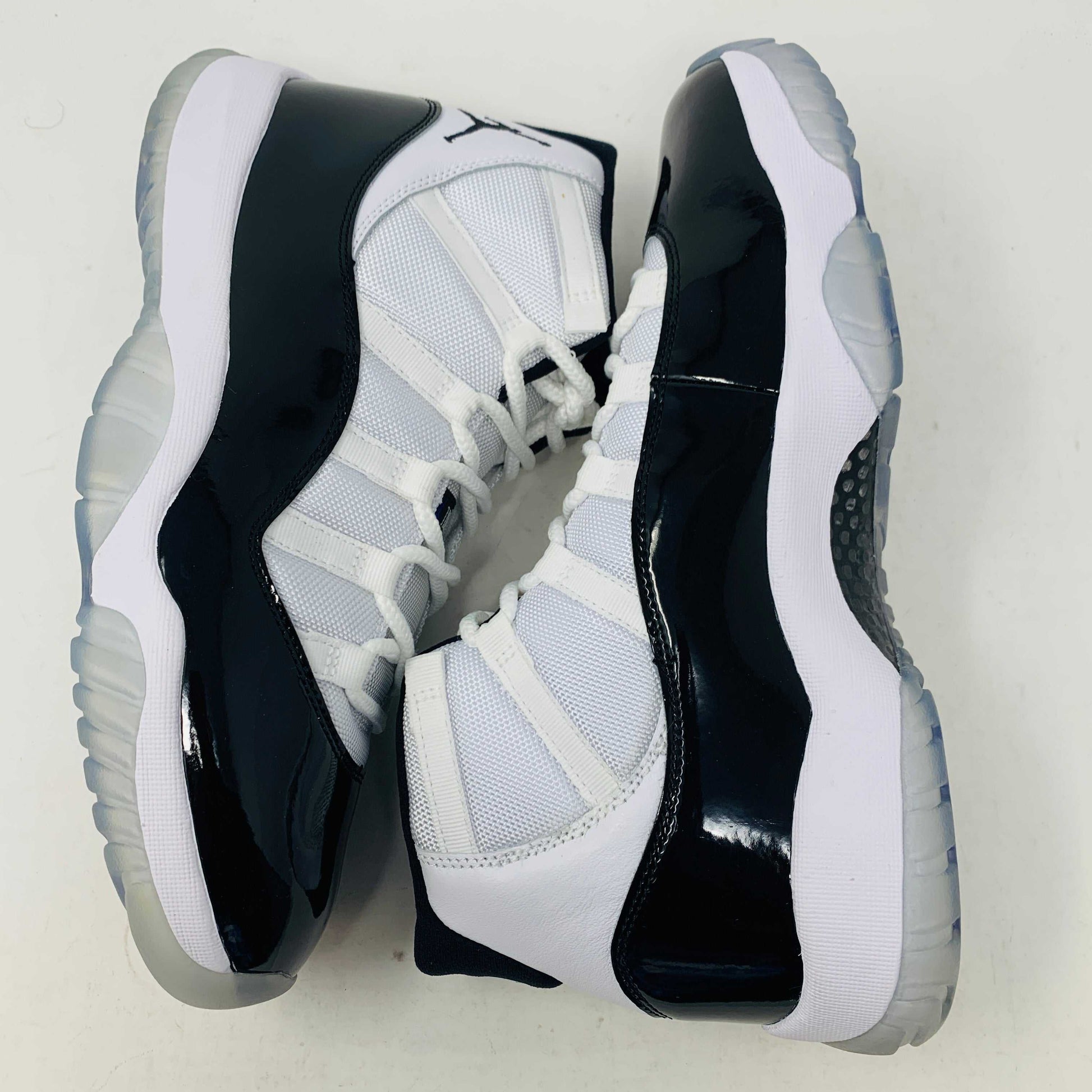 A BRAND NEW black and white Jordan 11 Concord 2018 sneaker sits on its shoebox, showcasing a glossy black patent leather finish and white mesh upper with the iconic Jumpman logo. The shoebox, bearing a white Nike Air logo, ensures these shoes are 100% authentic.
