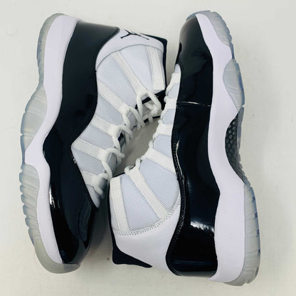 A BRAND NEW black and white Jordan 11 Concord 2018 sneaker sits on its shoebox, showcasing a glossy black patent leather finish and white mesh upper with the iconic Jumpman logo. The shoebox, bearing a white Nike Air logo, ensures these shoes are 100% authentic.