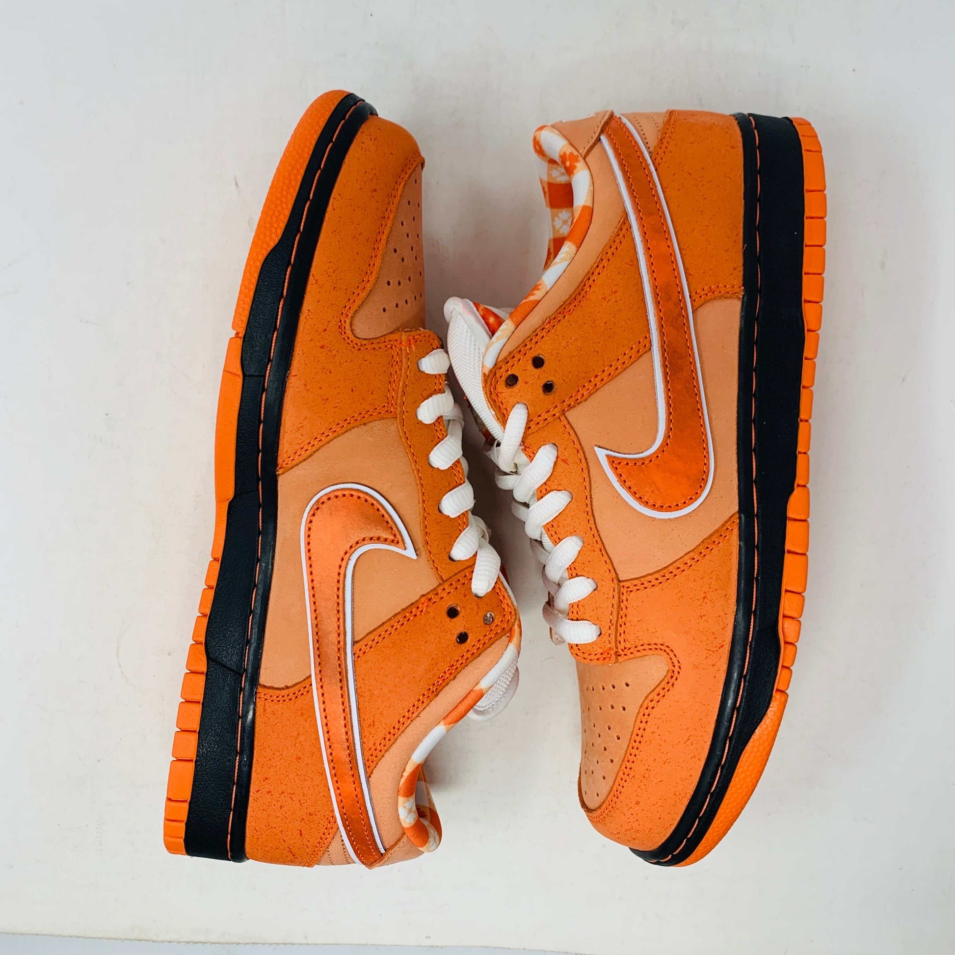 The authentic Nike SB Dunk Low Orange Lobster Concepts sneaker sits on a green and yellow Nike SB box. It features a white sole with black trim, an orange swoosh, and extra laces neatly positioned in front of the box.