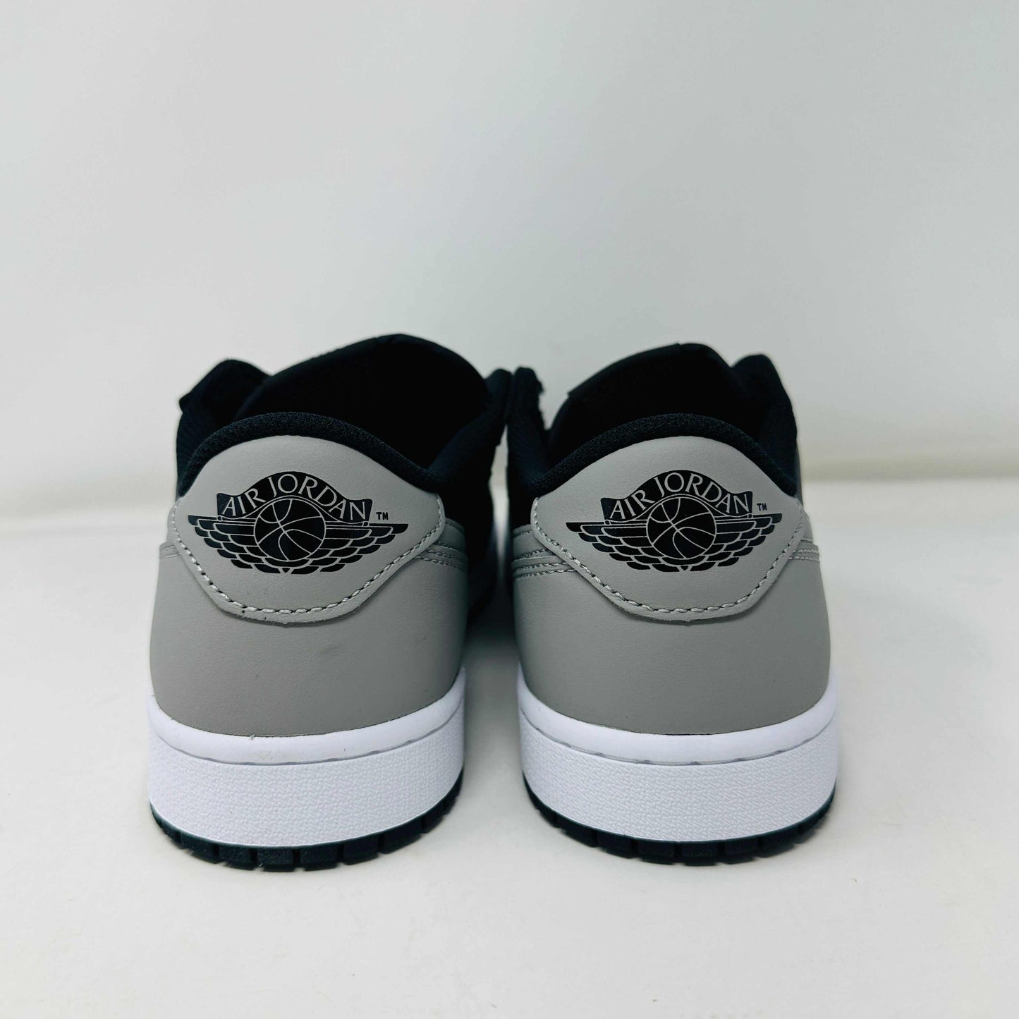 A pair of Jordan 1 Low OG Shadow (2024) sneakers, in black and gray, rests atop a gray shoe box with a large black Nike logo. Beside the box is a small brown cardboard package featuring a cutout revealing shoelaces inside.