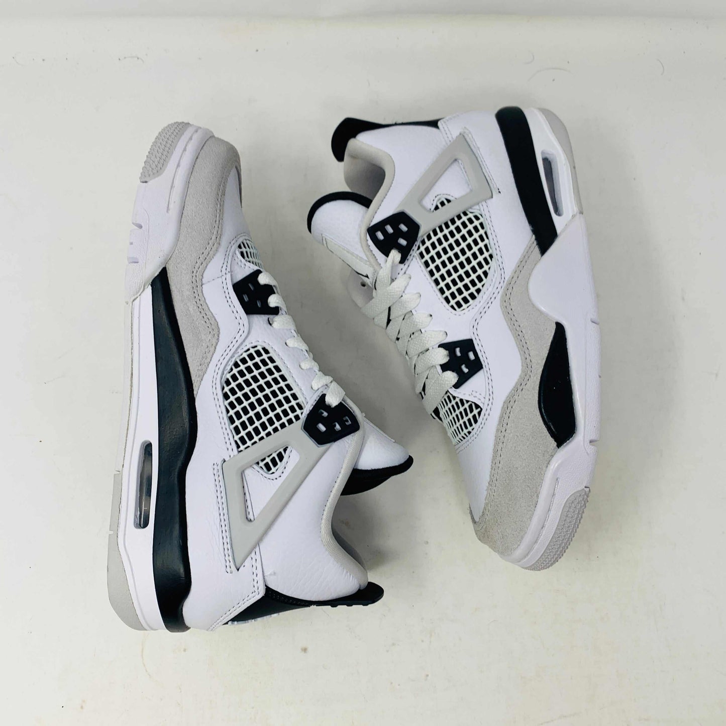 The Jordan 4 Military Black GS sneakers, featuring distinctive side netting and a logo on the tongue, are brand new and authentic in white and gray with black accents. They face forward on a white surface against a plain background, embodying modern style.