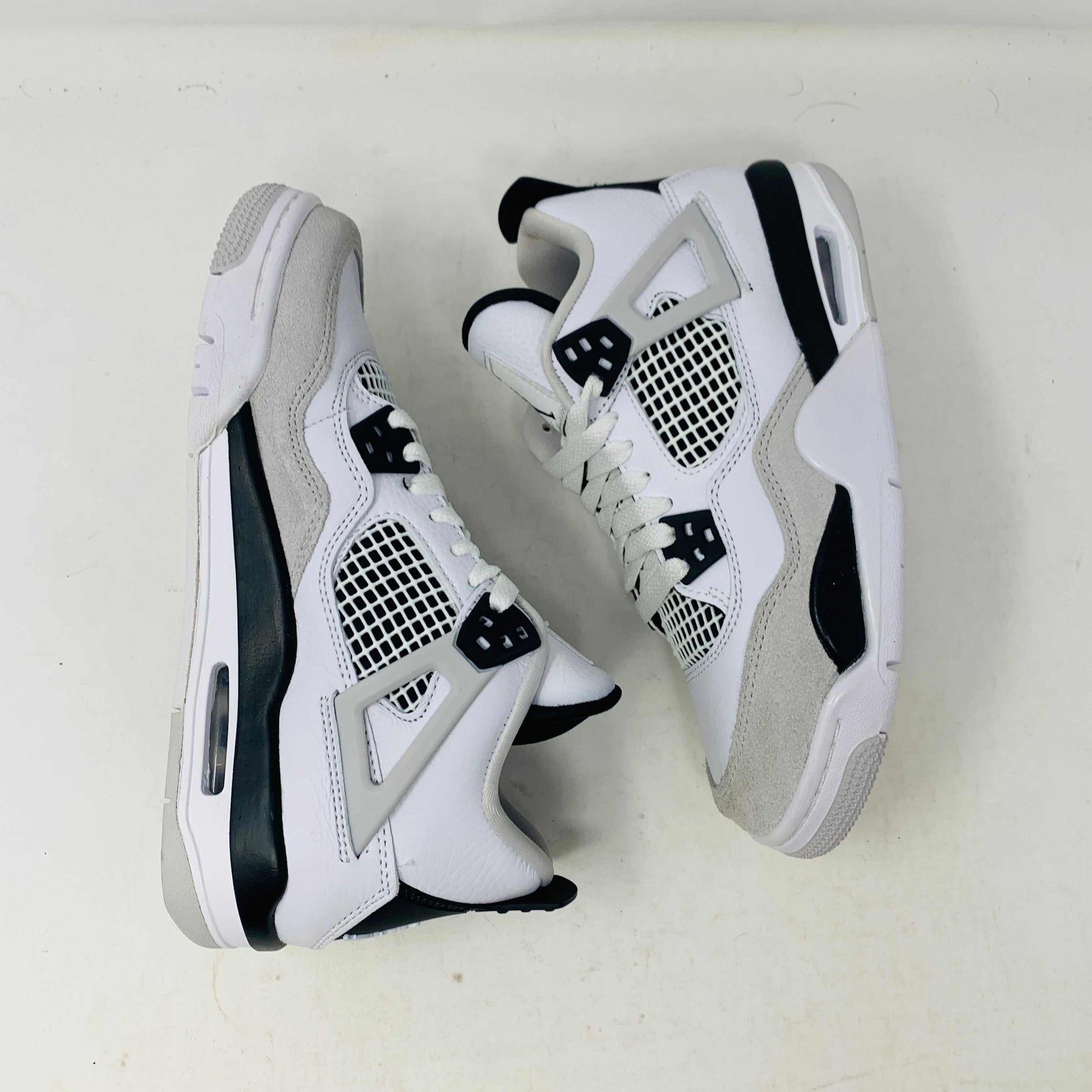 The Jordan 4 Military Black GS sneakers, featuring distinctive side netting and a logo on the tongue, are brand new and authentic in white and gray with black accents. They face forward on a white surface against a plain background, embodying modern style.