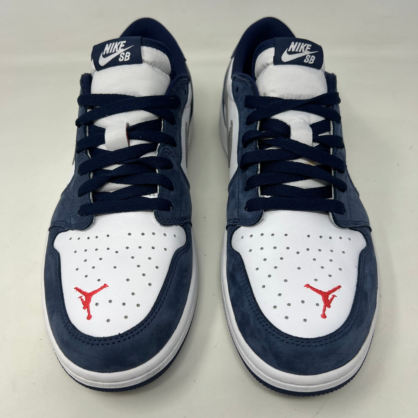 A Jordan 1 Low SB Midnight Navy shoe from Jordan, featuring blue toe and heel, white side panels, and a silver swoosh, is displayed on its box. The VERY CLEAN UPPERS are complemented by the size and product info visible on the label.
