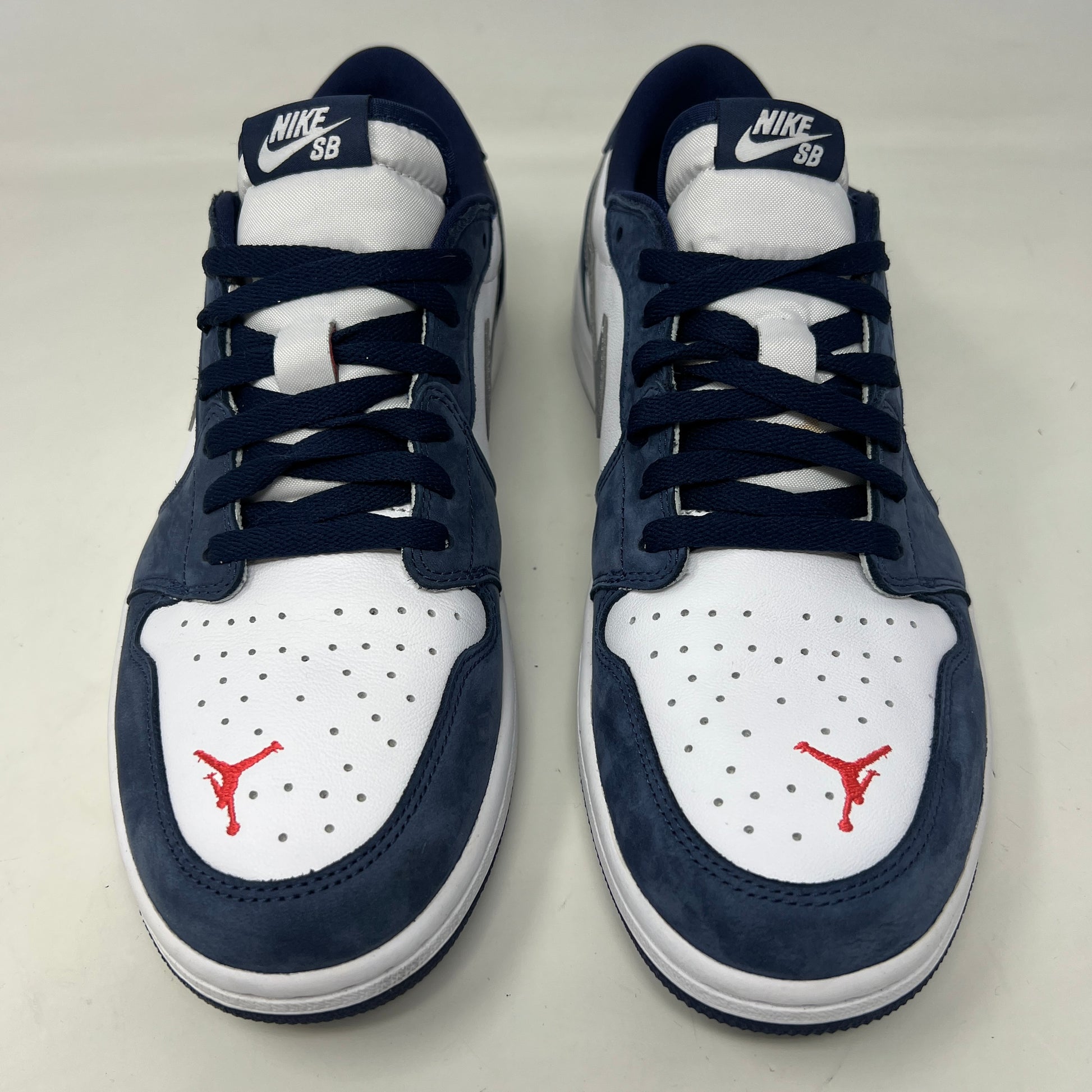 A Jordan 1 Low SB Midnight Navy shoe from Jordan, featuring blue toe and heel, white side panels, and a silver swoosh, is displayed on its box. The VERY CLEAN UPPERS are complemented by the size and product info visible on the label.