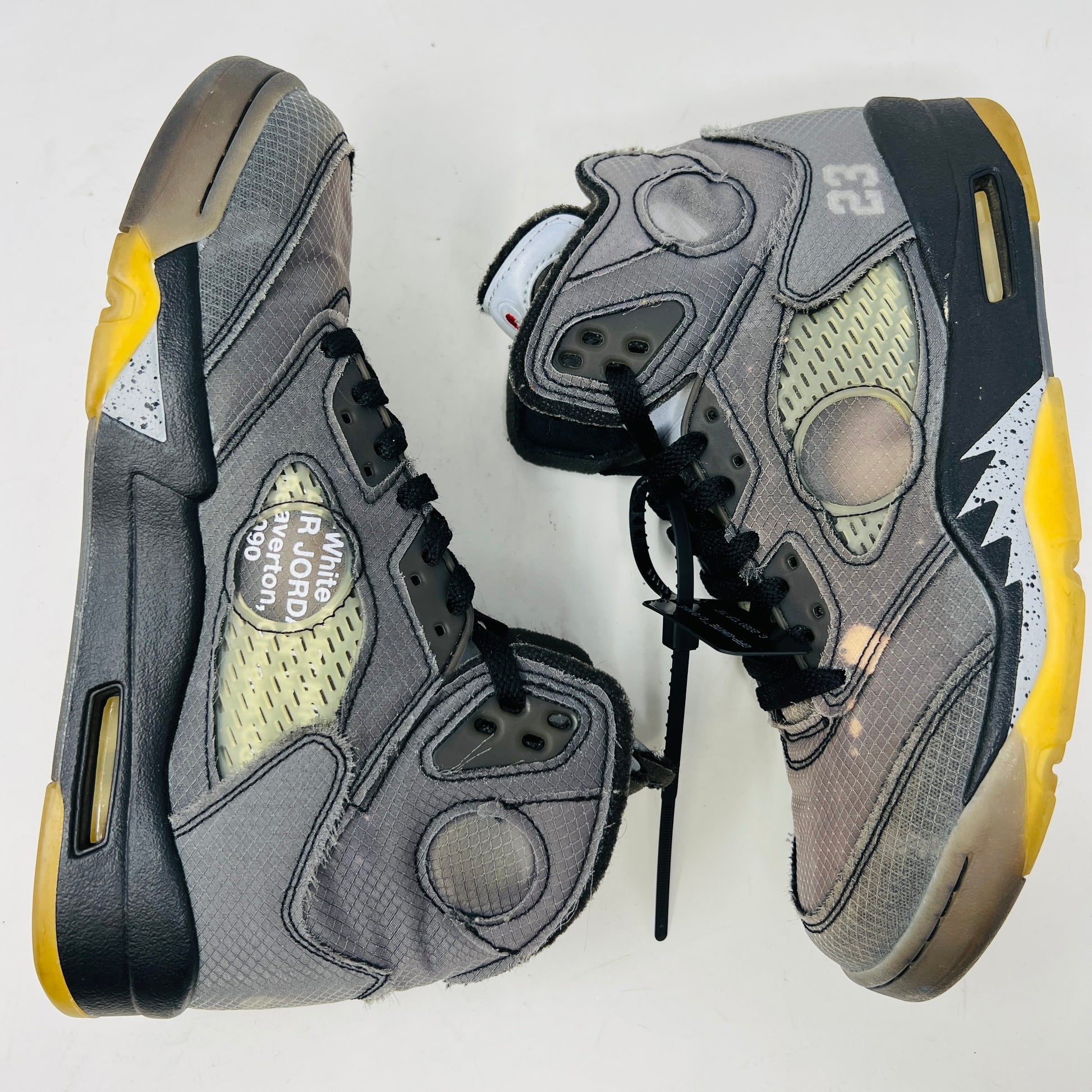 The Jordan 5 Retro Off-White Muslin high-top sneaker, featuring a transparent mesh window and 23 on the heel, is displayed on its box with a white and red shoelace packet in front. A small bleach stain adds unique character to the design.