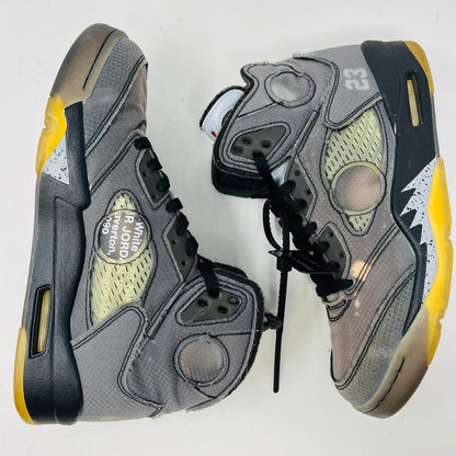 The Jordan 5 Retro Off-White Muslin high-top sneaker, featuring a transparent mesh window and 23 on the heel, is displayed on its box with a white and red shoelace packet in front. A small bleach stain adds unique character to the design.