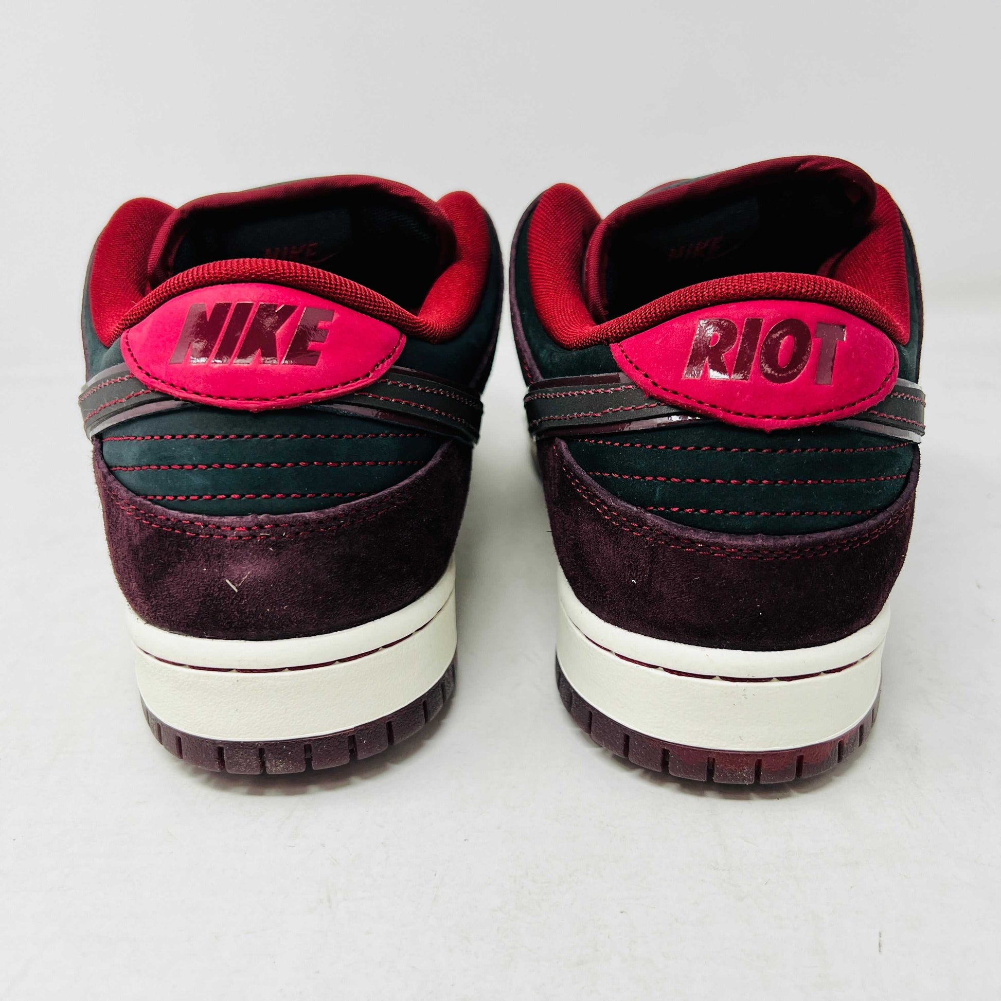 A BRAND NEW Nike SB Dunk Low Riot Skateshop sneaker featuring a black design with maroon accents, including a swoosh, laces, and a black sole with a white midsole, is displayed on top of its original white Nike SB shoebox.