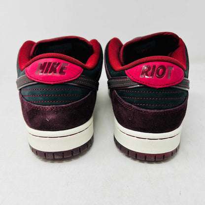 A BRAND NEW Nike SB Dunk Low Riot Skateshop sneaker featuring a black design with maroon accents, including a swoosh, laces, and a black sole with a white midsole, is displayed on top of its original white Nike SB shoebox.