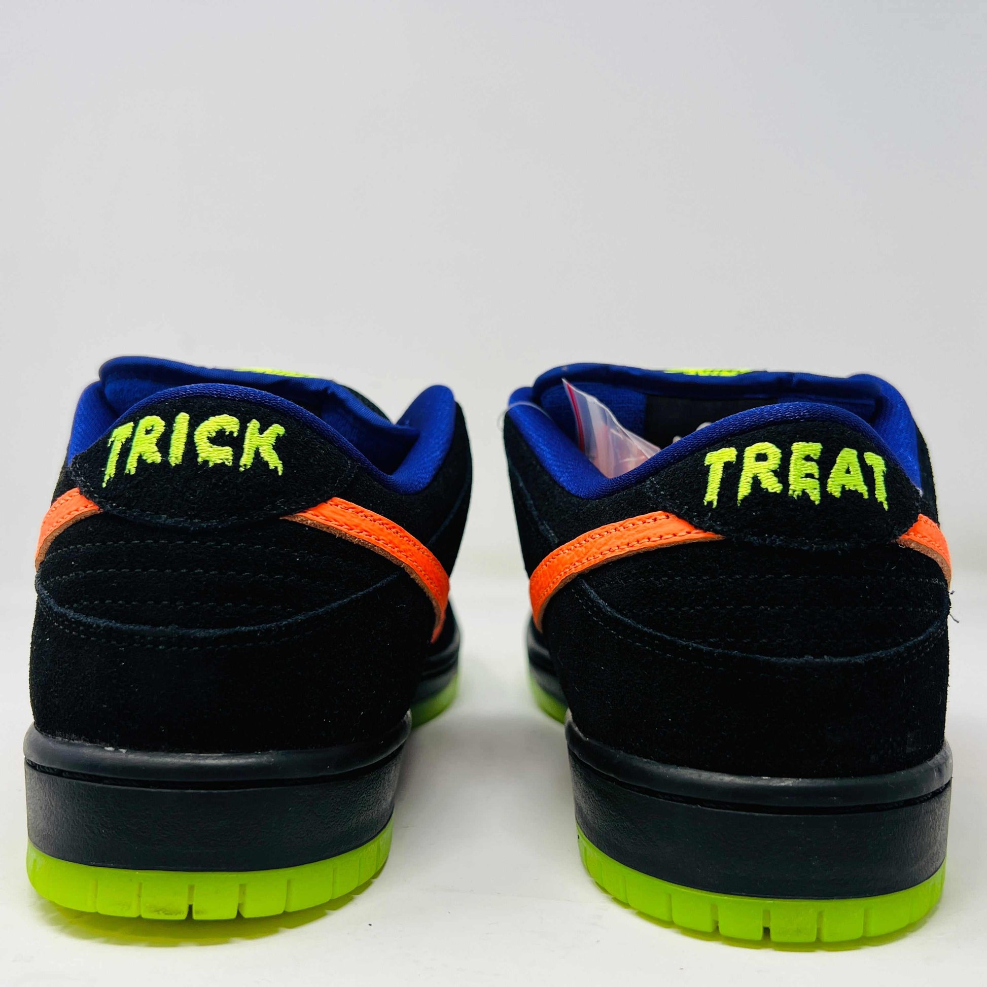 The Nike SB Dunk Low Night of Mischief sneakers feature Halloween-themed designs with orange swooshes, green soles, and spider web details. They come in a coffin-shaped box adorned with spooky jack-o-lanterns and webs, resting on a wooden floor.