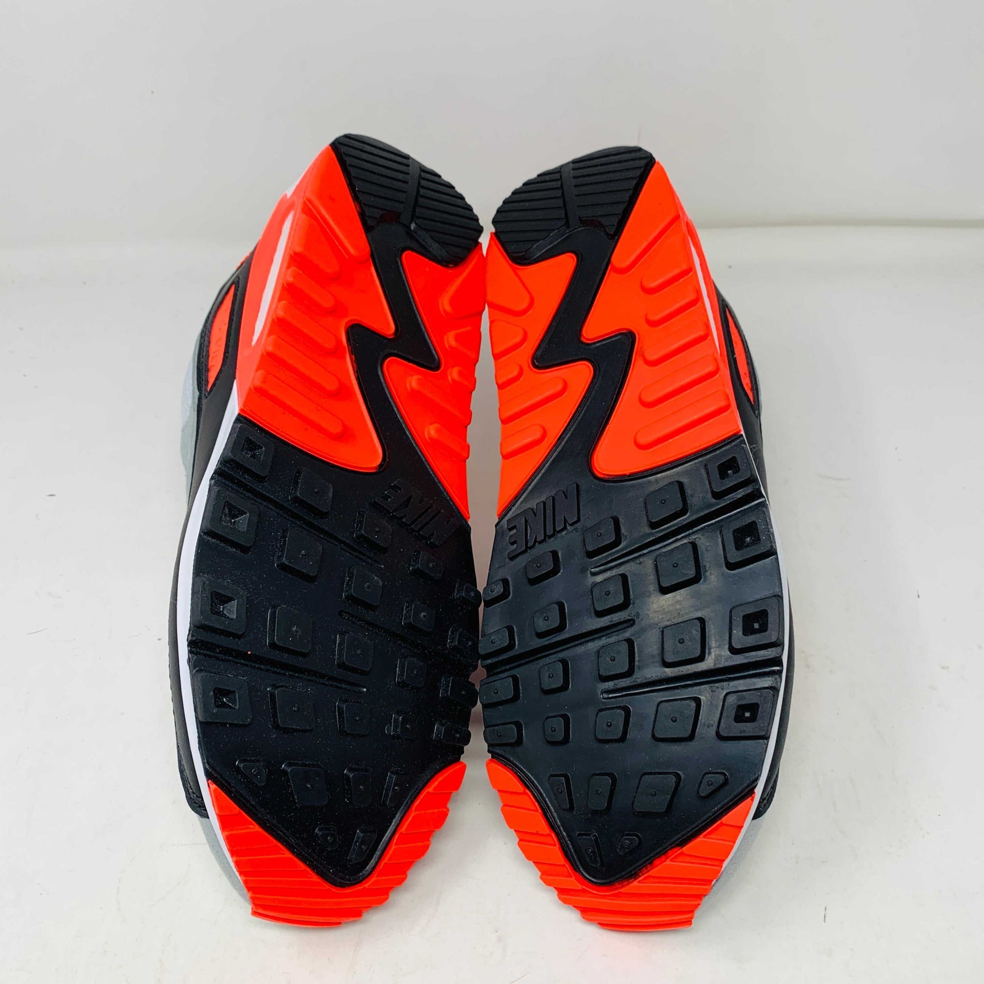 A pair of Nike Air Max 90 Infrared (2020) sneakers in gray, white, and bright orange sits on a pristine shoebox. Beside the shoes are stylish accessories: an orange keychain and spare orange soles.