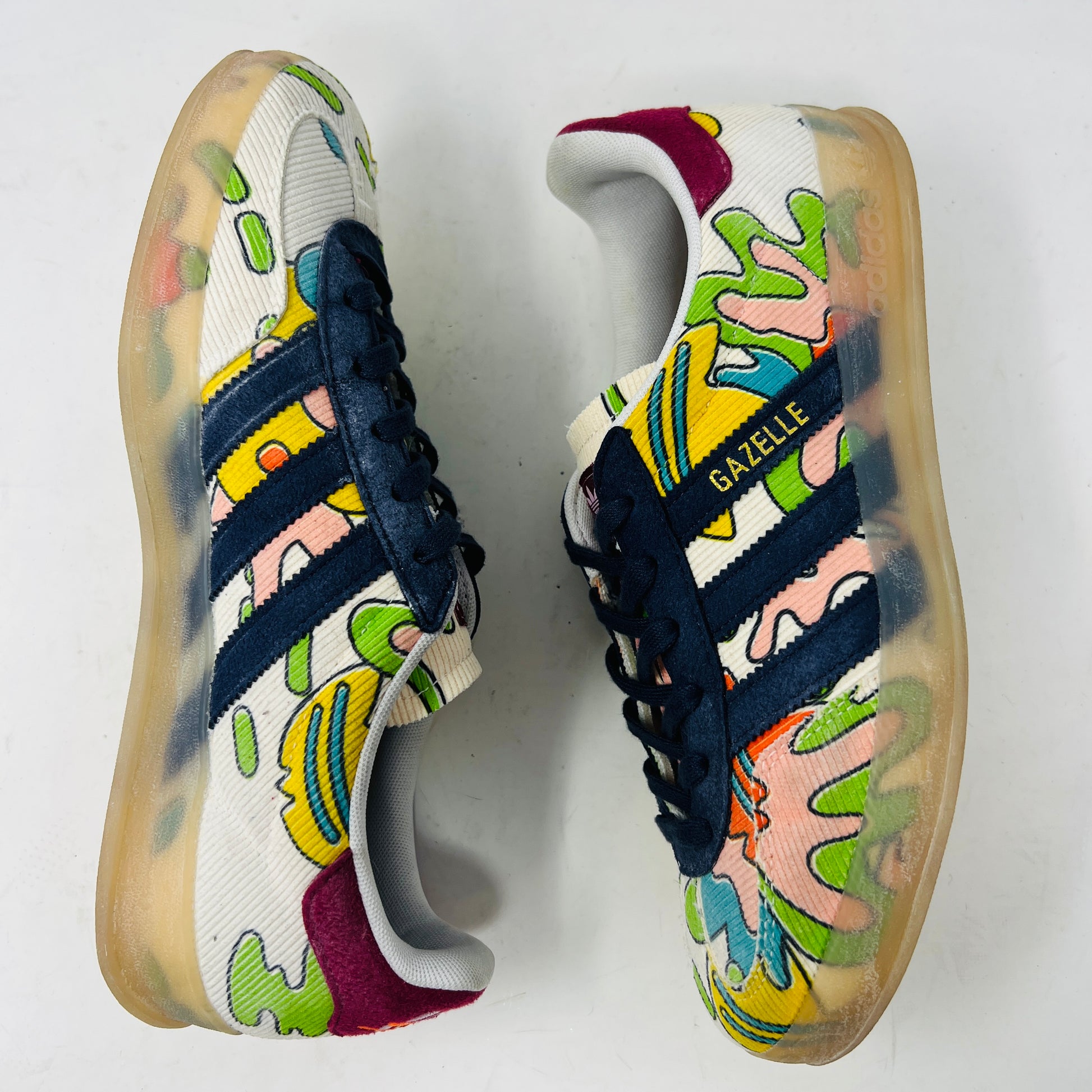 The Adidas Gazelle Indoor Sean Wotherspoon sneaker, with colorful abstract uppers in yellow, red, pink, and green, dark blue stripes, red laces, and translucent soles, rests on a blue box showing size and product details.
