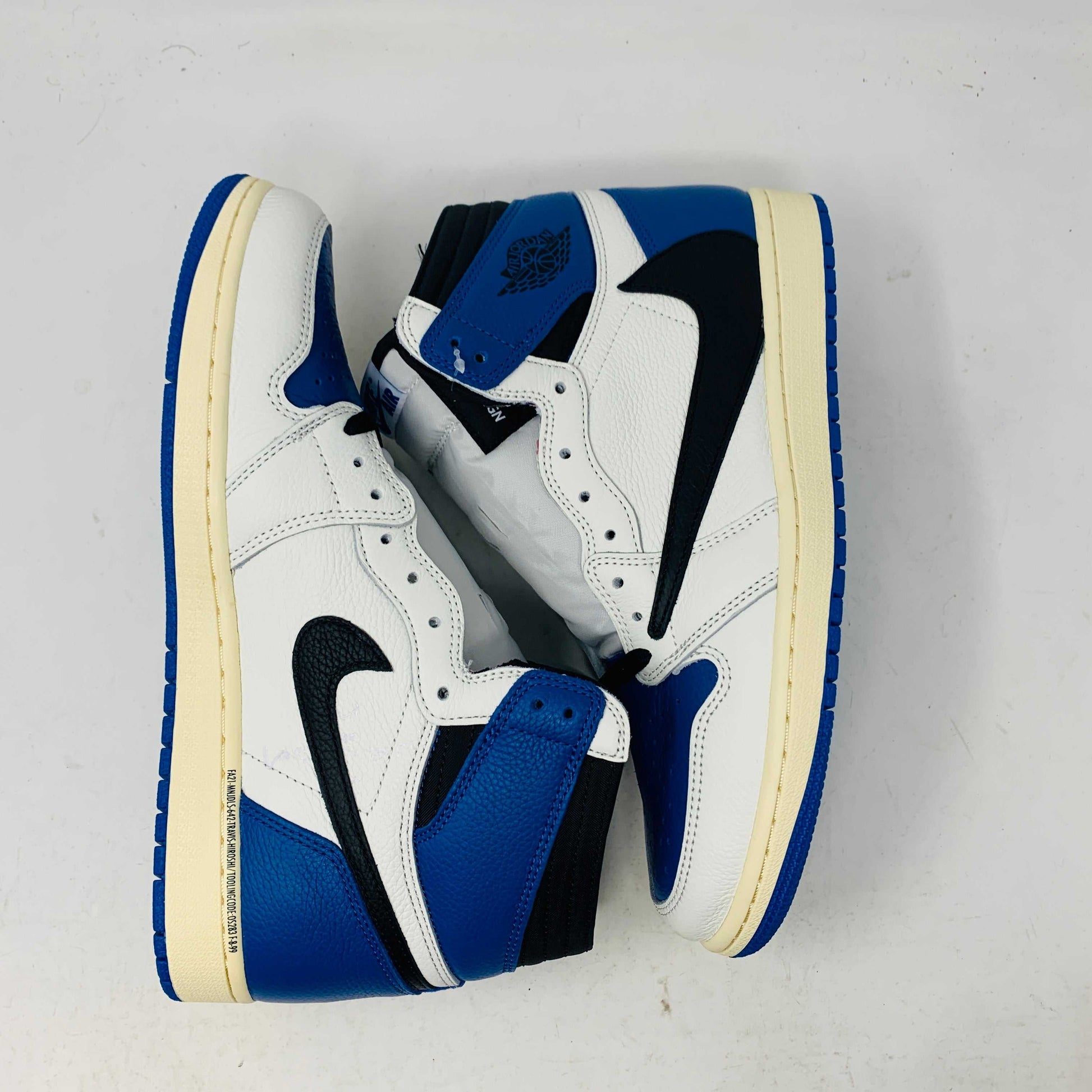 The Jordan 1 Retro High OG SP Fragment x Travis Scott is a new high-top sneaker featuring a blue, white, and black design with a unique backward Nike swoosh. It comes in a transparent, branded box adorned with butterfly and text details, boasting a thick sole and classic 1985 Air Jordan design.