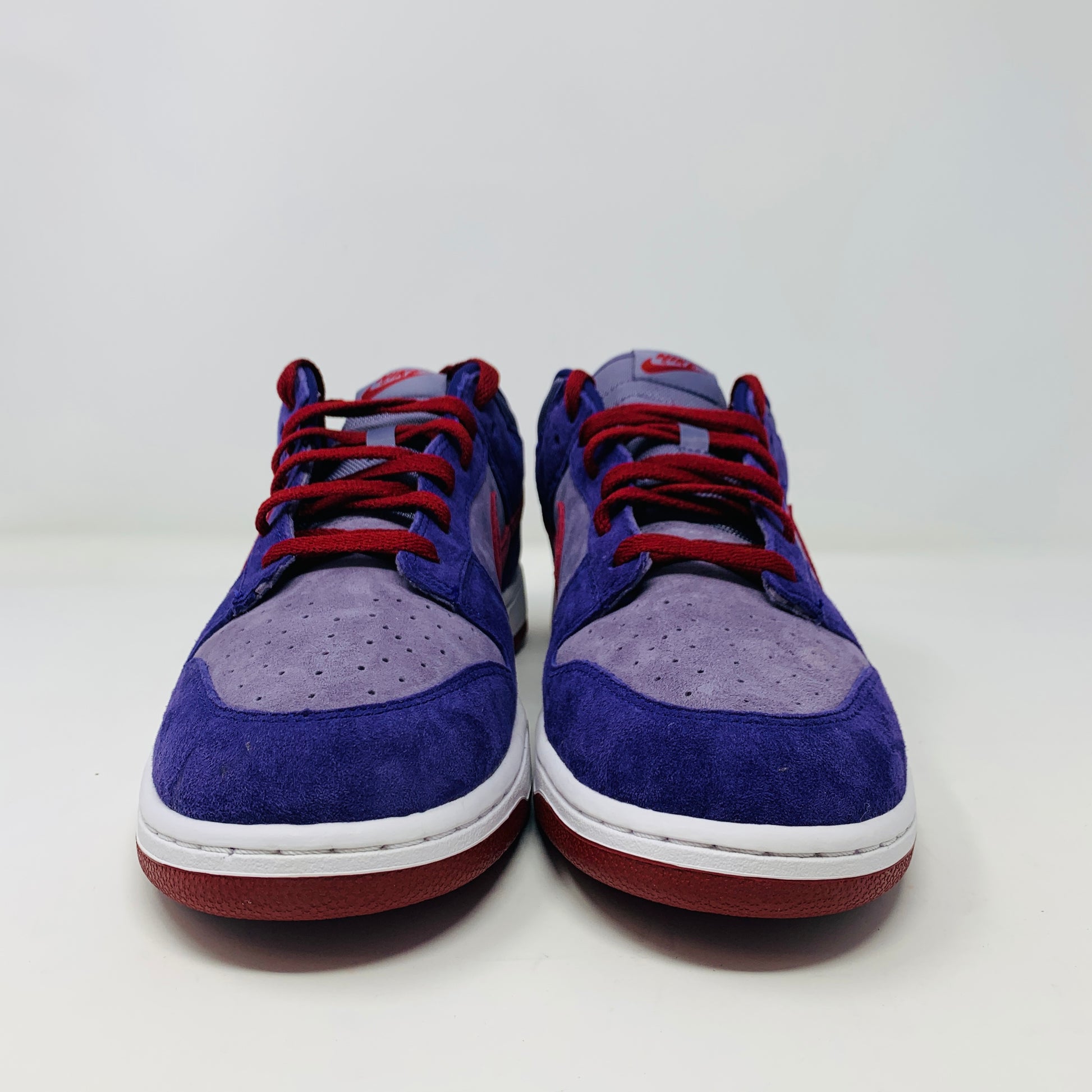 A pair of Nike Dunk Low Plum sneakers, in purple and maroon, rests on a red shoe box featuring a white Nike logo on a plain background. The shoes showcase a red swoosh and white midsoles, confirming their authenticity from Nike.