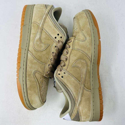The Nike SB Dunk Low Pro B Parachute Beige, with a beige swoosh and gum sole, is displayed on a purple Nike SB box. New tan suede sneaker includes extra laces neatly rolled up in front against a plain white background.