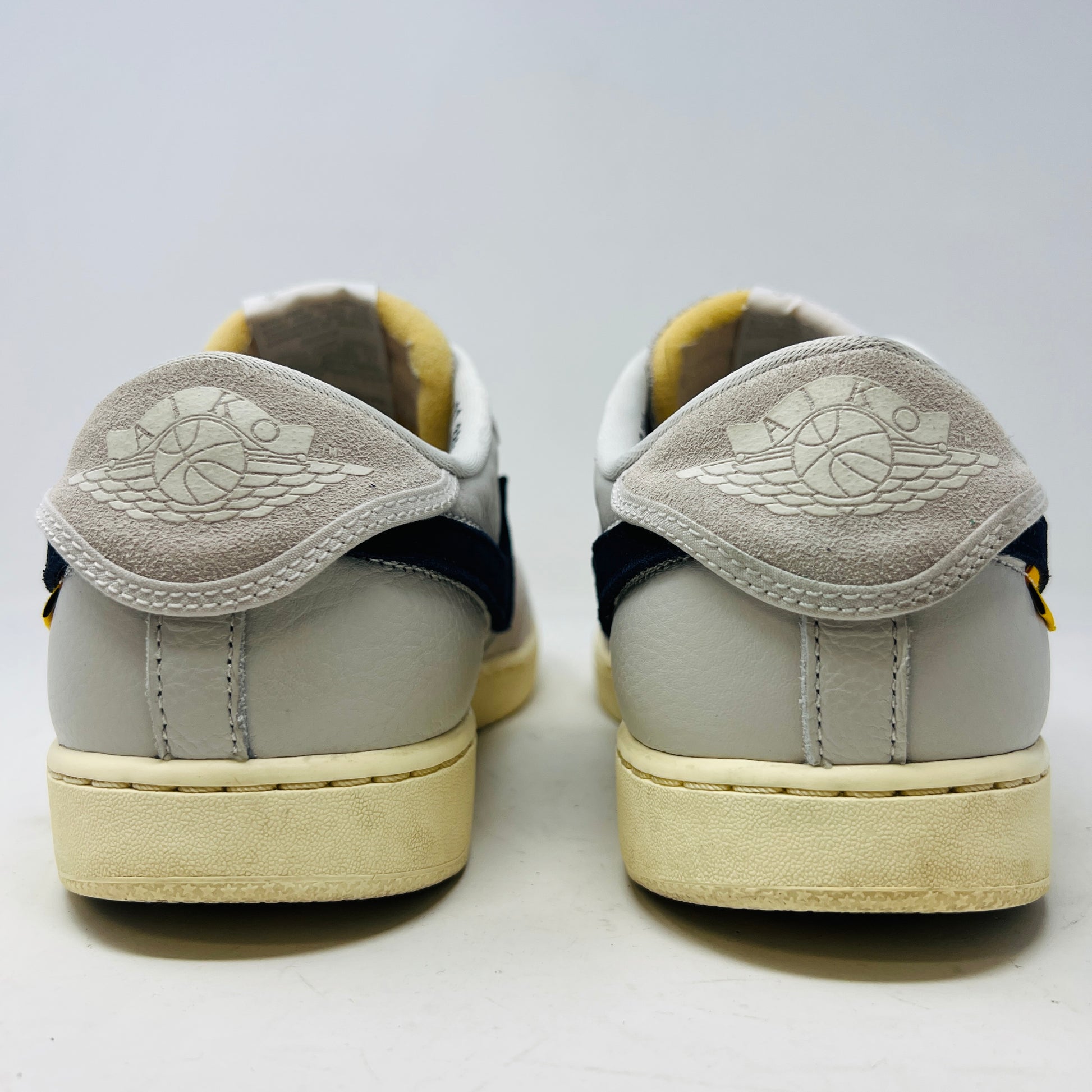 The Jordan 1 Retro AJKO Low SP Union Sail Leather sneakers feature black swooshes, subtle yellow stitching, and white laces. Pictured on a pristine white background, they include extra laces for versatile styling.