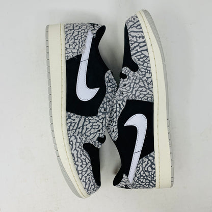 The Jordan 1 Retro Low OG Black Cement sneaker, with black and gray elephant print and a white swoosh, is showcased on a matching shoe box. A small brown box with extra laces is beside it, all set against a plain background.