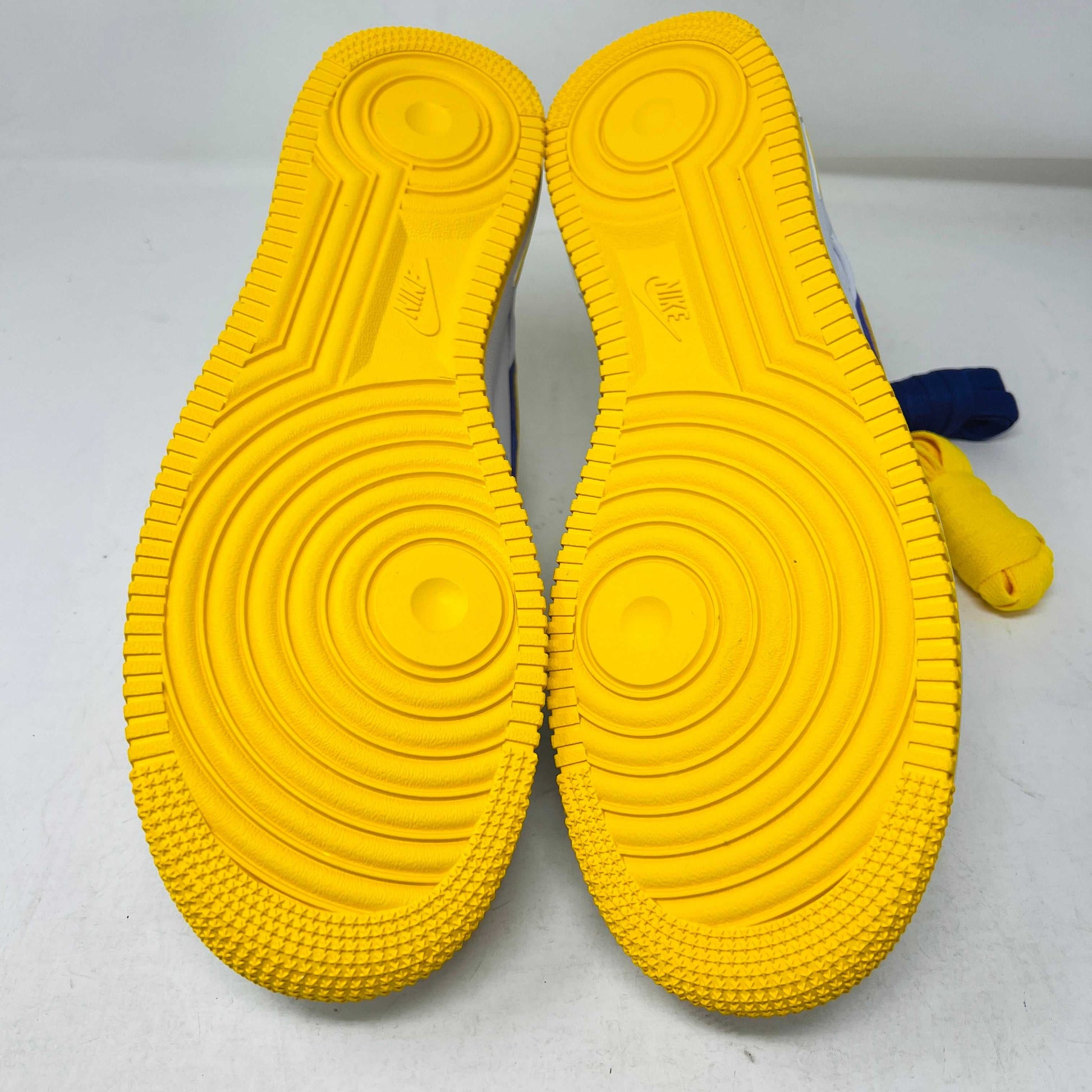 The Nike Air Force 1 Low Retro QS Kobe Bryant Lakers Home sneakers showcase their bright yellow soles with intricate tread patterns, featuring circular grooves and textured edges, positioned soles up.
