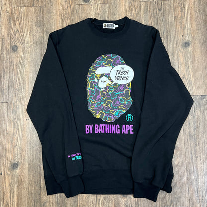This pre-owned black A BATHING APE X THE FRESH PRINCE sweatshirt by Bape features a vibrant ape graphic with sunglasses, The Fresh Prince in a speech bubble, and purple and green logo accents, all displayed on a wooden floor.