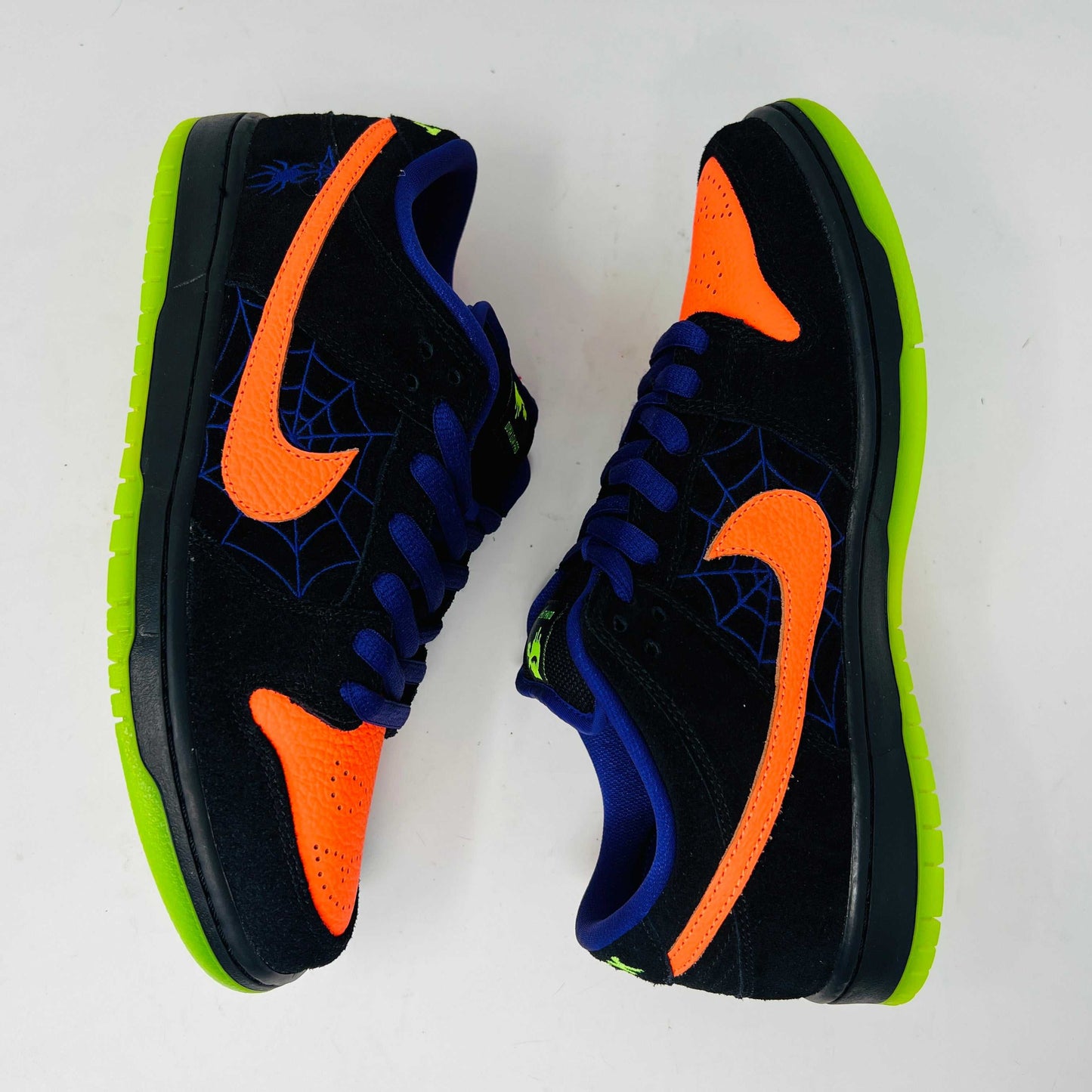The Nike SB Dunk Low Night of Mischief sneakers feature Halloween-themed designs with orange swooshes, green soles, and spider web details. They come in a coffin-shaped box adorned with spooky jack-o-lanterns and webs, resting on a wooden floor.
