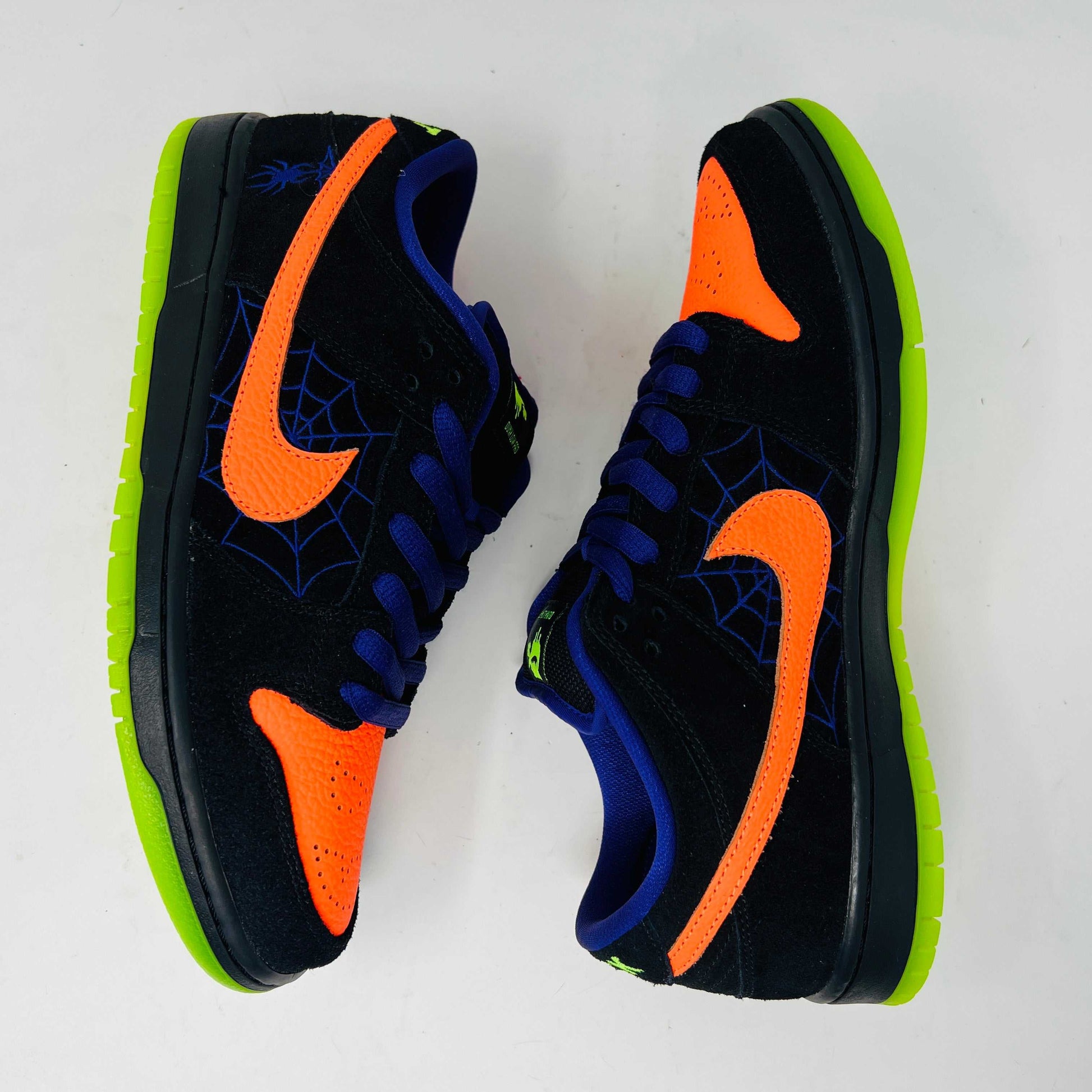 The Nike SB Dunk Low Night of Mischief sneakers feature Halloween-themed designs with orange swooshes, green soles, and spider web details. They come in a coffin-shaped box adorned with spooky jack-o-lanterns and webs, resting on a wooden floor.