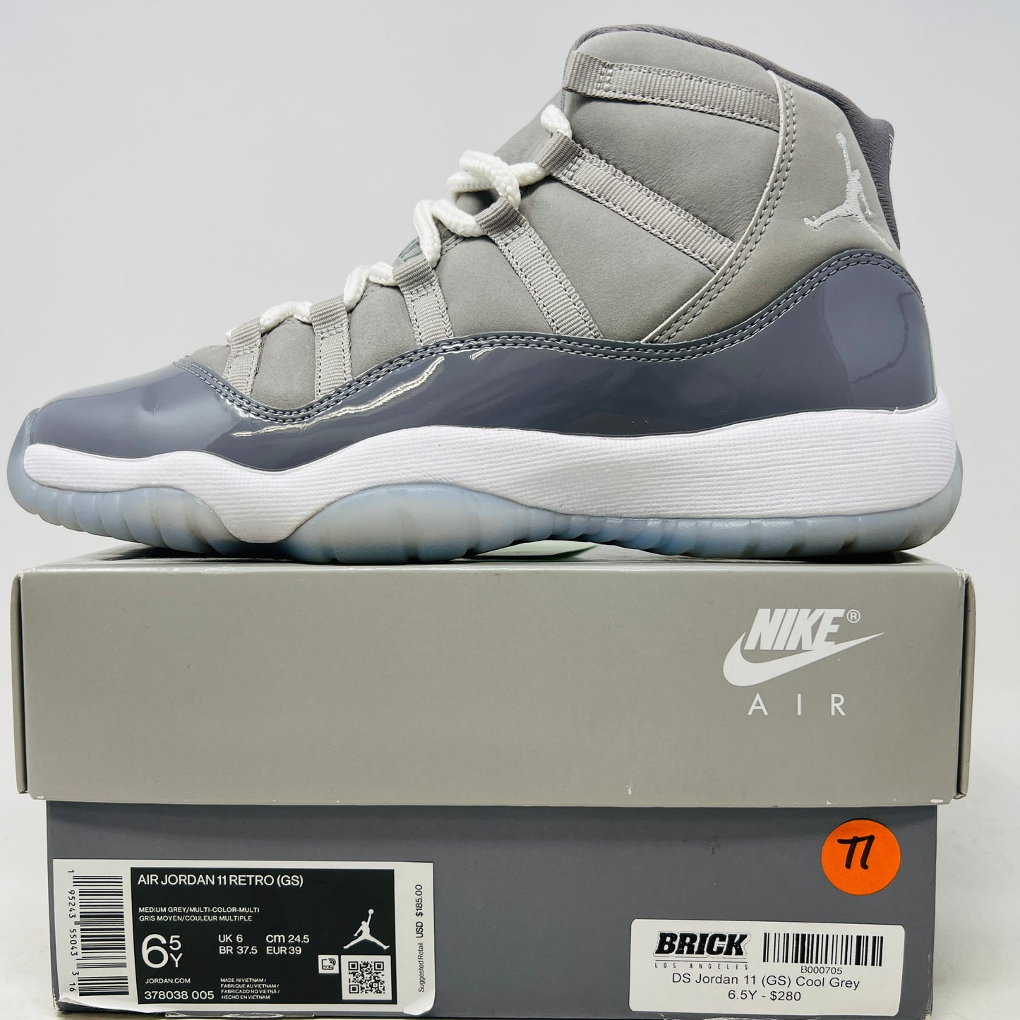 A Jordan 11 Retro Cool Grey (2021) (GS) sneaker by Jordan, in gray and white with a glossy patent leather finish, is displayed on its original Nike shoebox. The box is in good condition featuring a size 6.5Y label and various details, with clean uppers showing light wear and an orange sticker numbered 71.