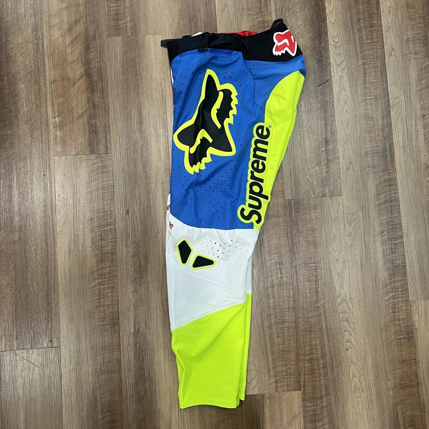 Supreme Fox Racing Moto Pant Multicolor features vibrant blue, yellow, and white panels with Supreme printed vertically on each leg. Brand new with black waist trim and fox logos on the thighs, they are displayed on a wooden floor.