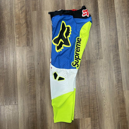 Supreme Fox Racing Moto Pant Multicolor features vibrant blue, yellow, and white panels with Supreme printed vertically on each leg. Brand new with black waist trim and fox logos on the thighs, they are displayed on a wooden floor.