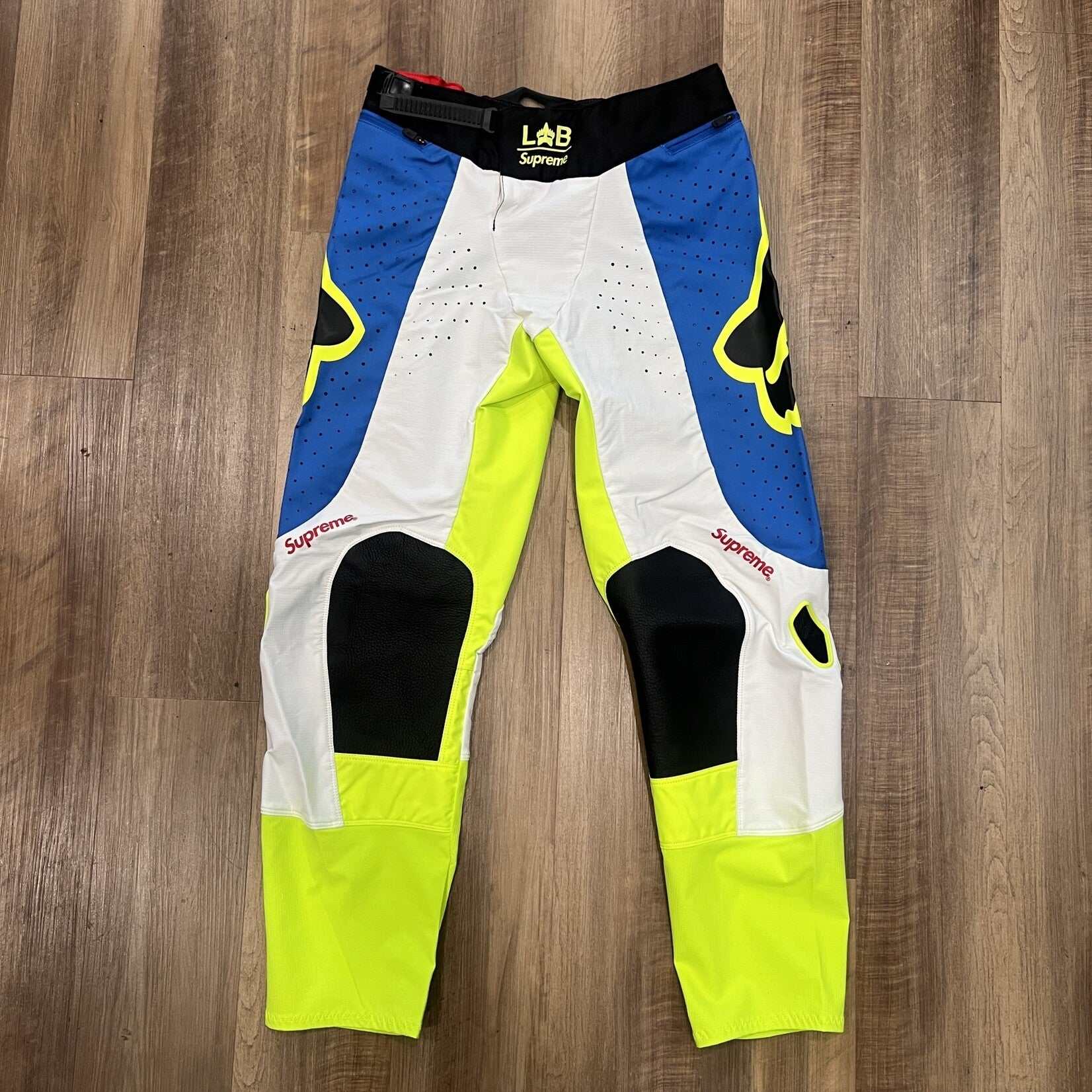 Supreme Fox Racing Moto Pant Multicolor features vibrant blue, yellow, and white panels with Supreme printed vertically on each leg. Brand new with black waist trim and fox logos on the thighs, they are displayed on a wooden floor.